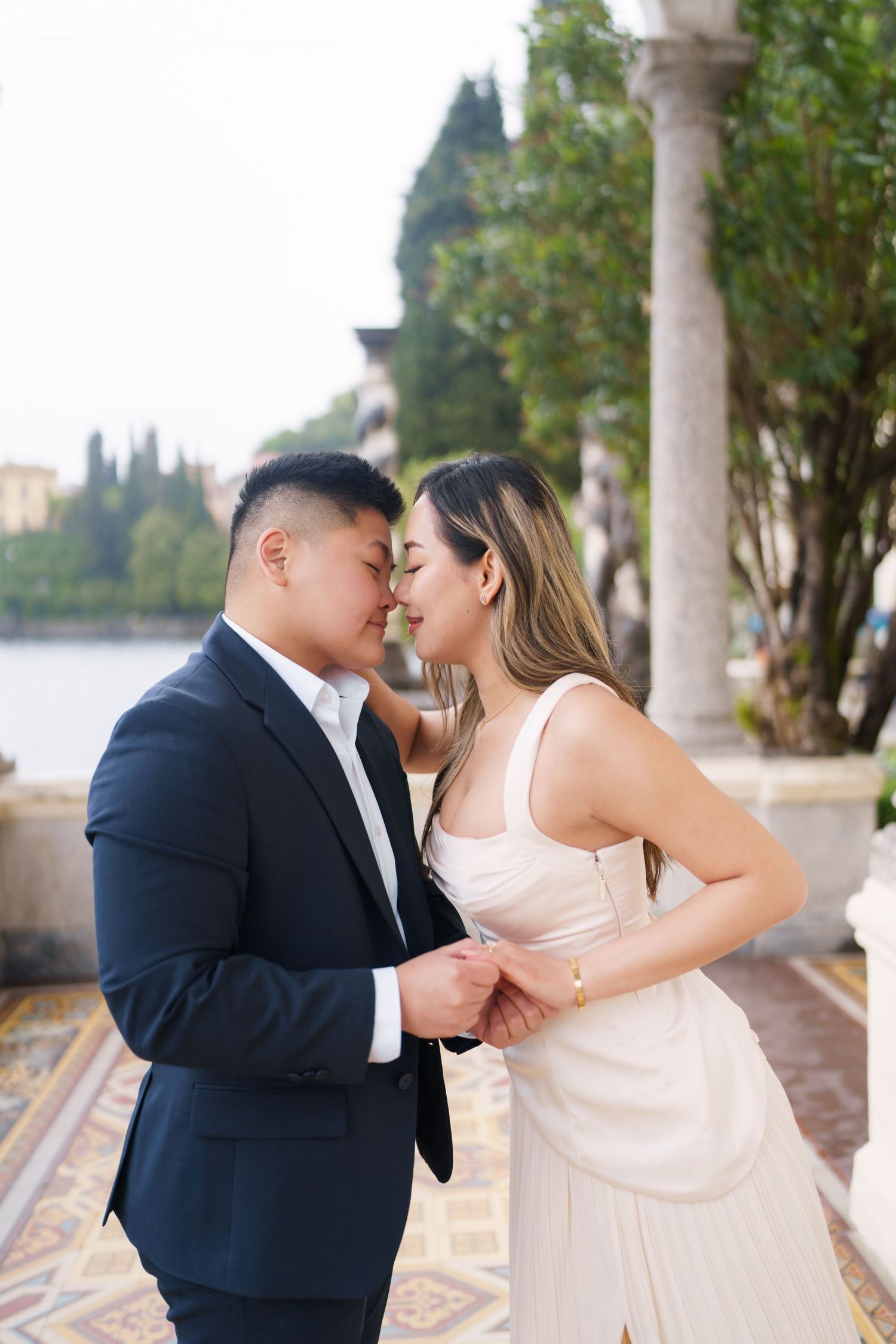 Couple in elegant attire sharing a romantic moment near a lakeside colonnade, man in navy suit, woman in ivory dress.