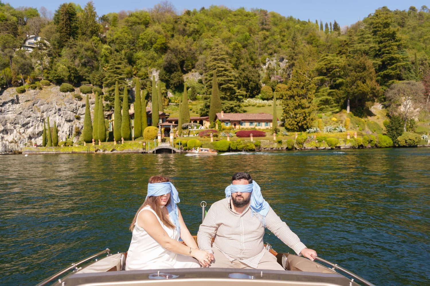 Couple wearing blue masks ride a boat on a lake with lush green trees and a red house in the background.