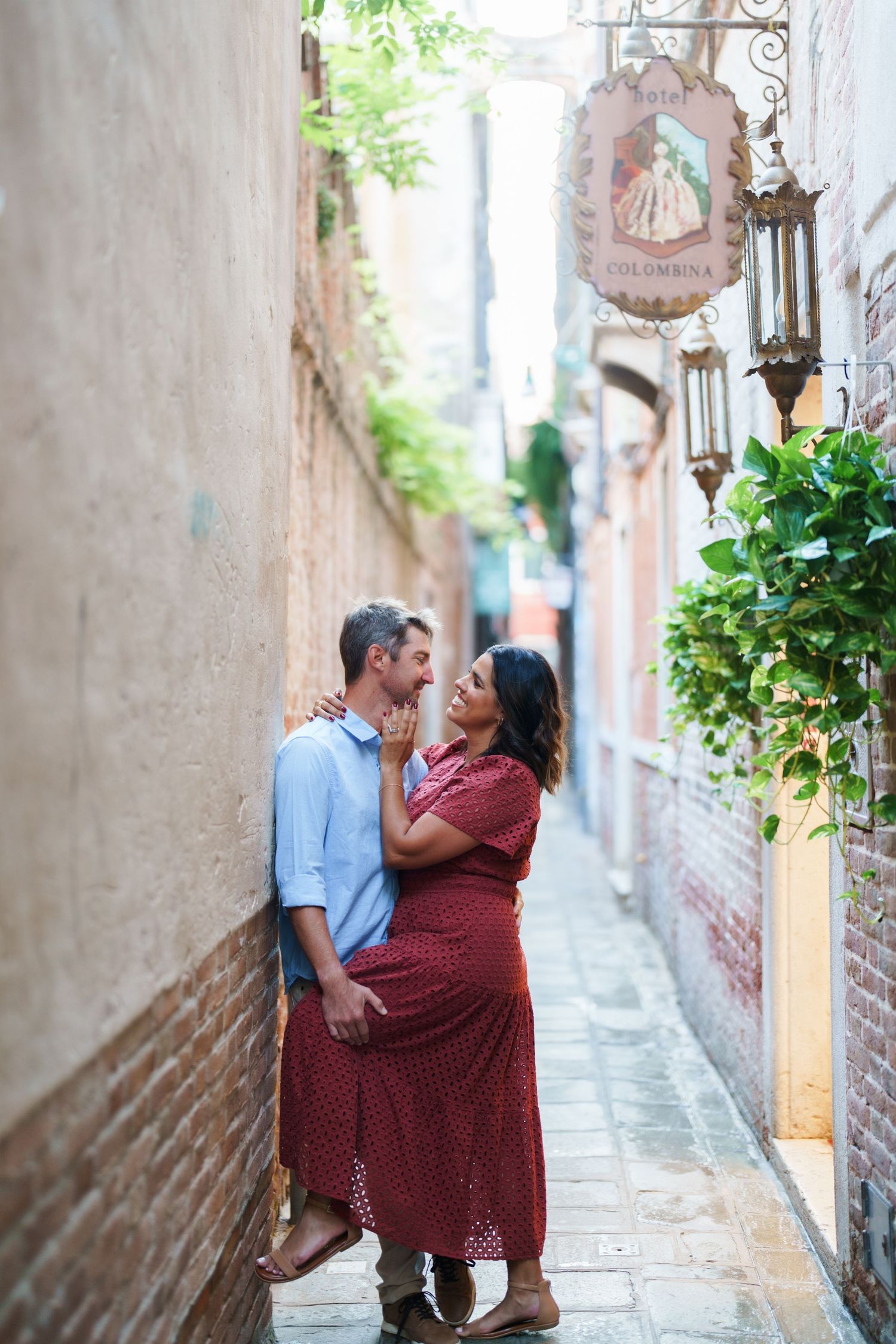 Couple embracing romantically in a narrow European alley beside brick walls and a decorative shop sign.