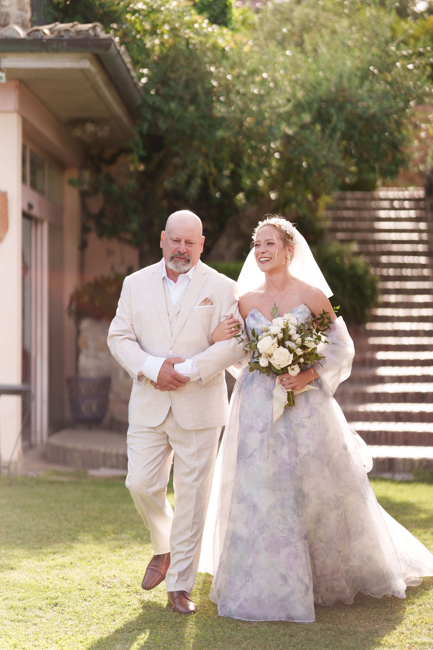 Bride in floral gown holding bouquet walks with man in beige suit through sunlit garden on wedding day.