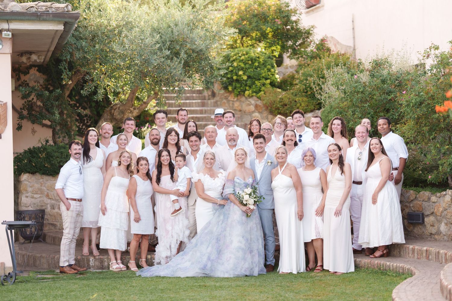 Large wedding party in white and gray attire poses outdoors on green lawn with stone steps and lush trees behind them.