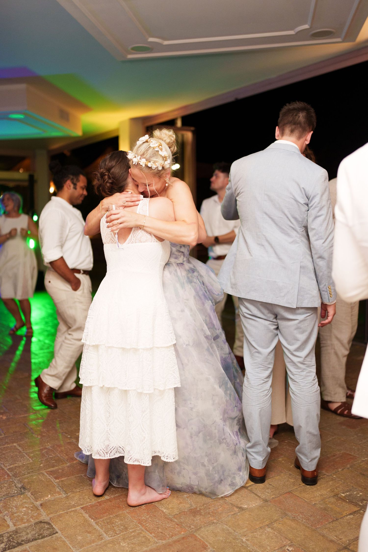 Bride and guest share a heartfelt hug on the dance floor at a wedding reception with colorful lighting.