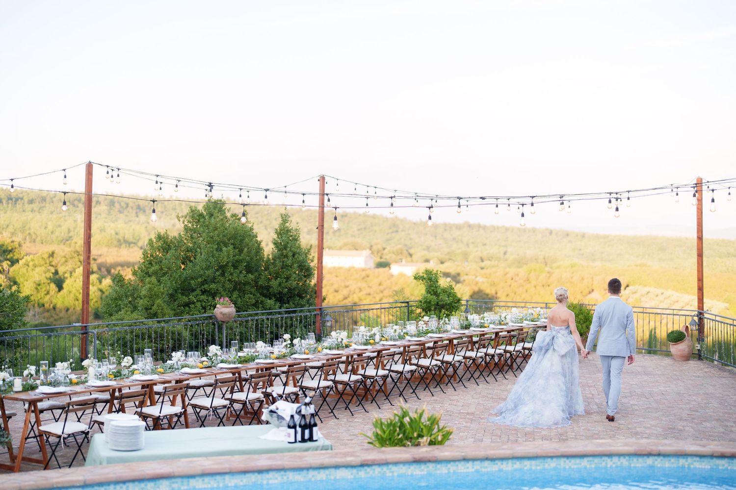 Bride and groom walk hand-in-hand toward a long outdoor reception table with string lights at a vineyard wedding.