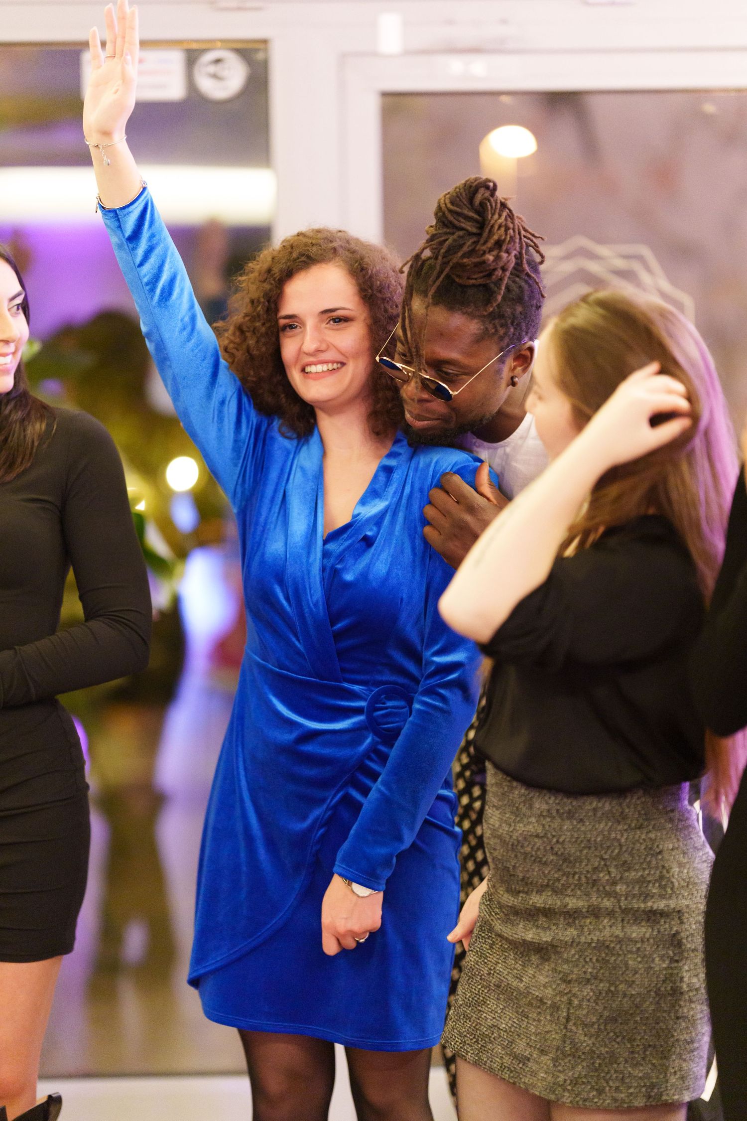 A woman in a blue velvet dress dancing with friends at a party, raising her hand joyfully.