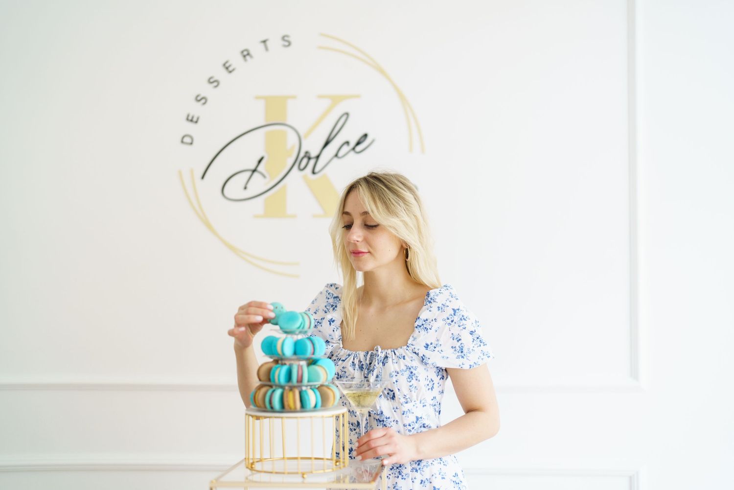 Blonde woman in floral dress arranging macarons on gold stand at Dolce Desserts bakery.