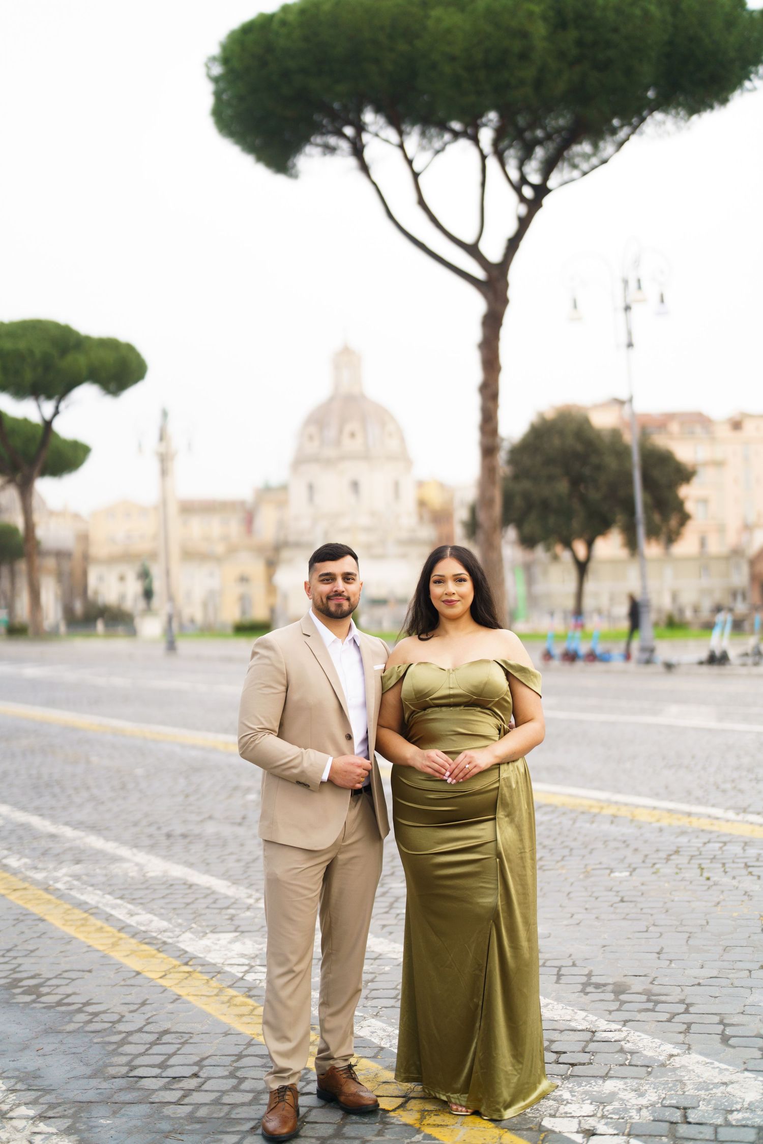 Elegant couple posing in Rome, man in beige suit and woman in olive green gown, with historic architecture backdrop.