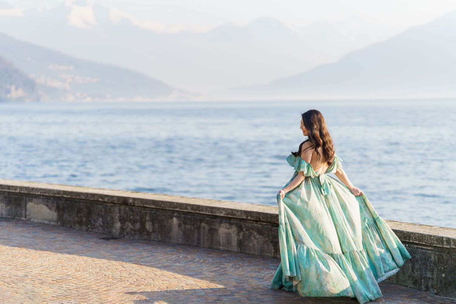 Woman in elegant teal gown standing by a stone promenade overlooking a serene lake with misty mountains.
