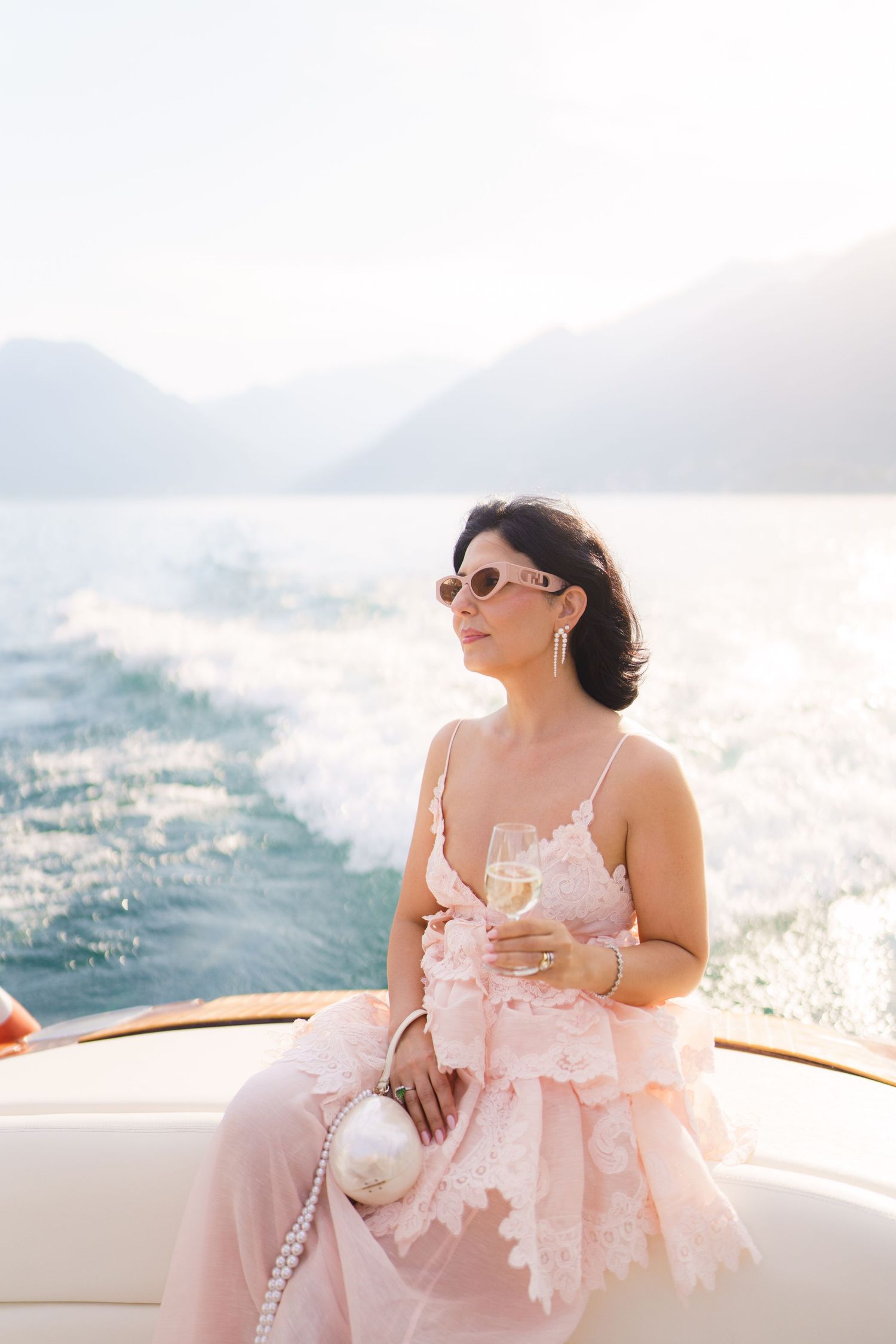 Elegant woman in pink lace dress holding champagne on a luxury yacht with mountain lake backdrop.