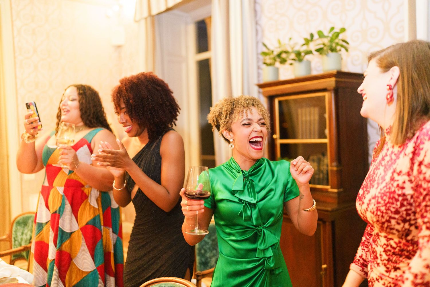 Women laughing and dancing at an indoor party, one in a green dress holding a wine glass.
