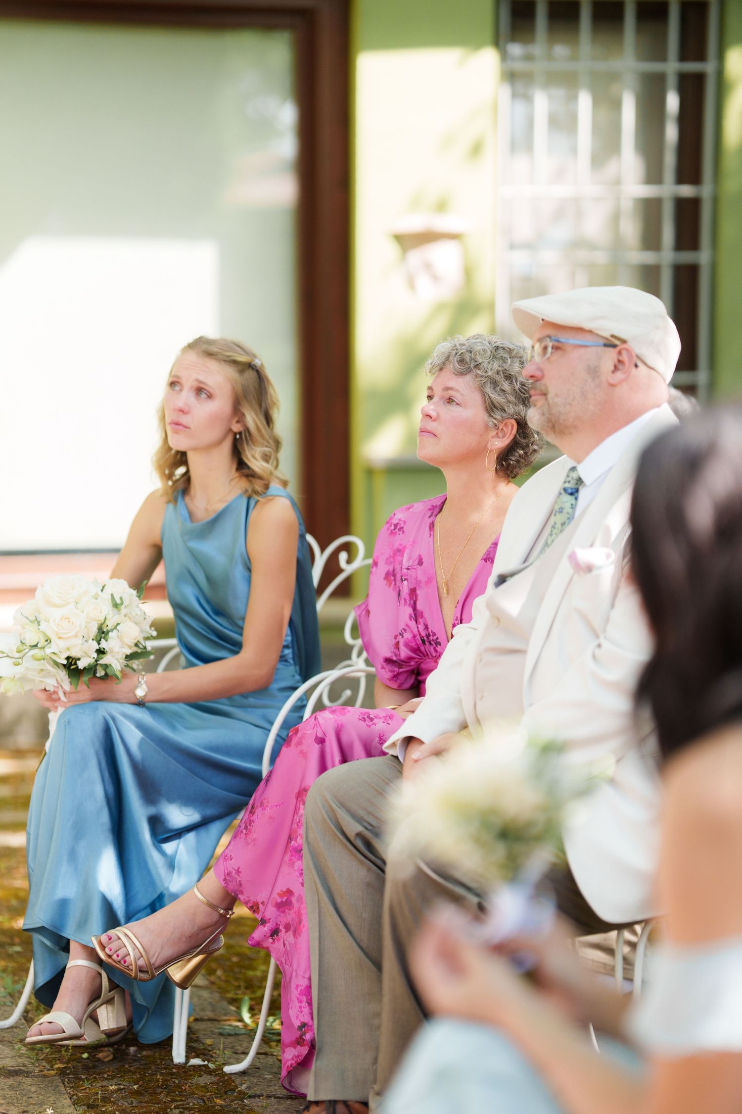 Wedding guests seated outdoors, including a bridesmaid in blue dress holding white flowers, and an older couple in formal attire.