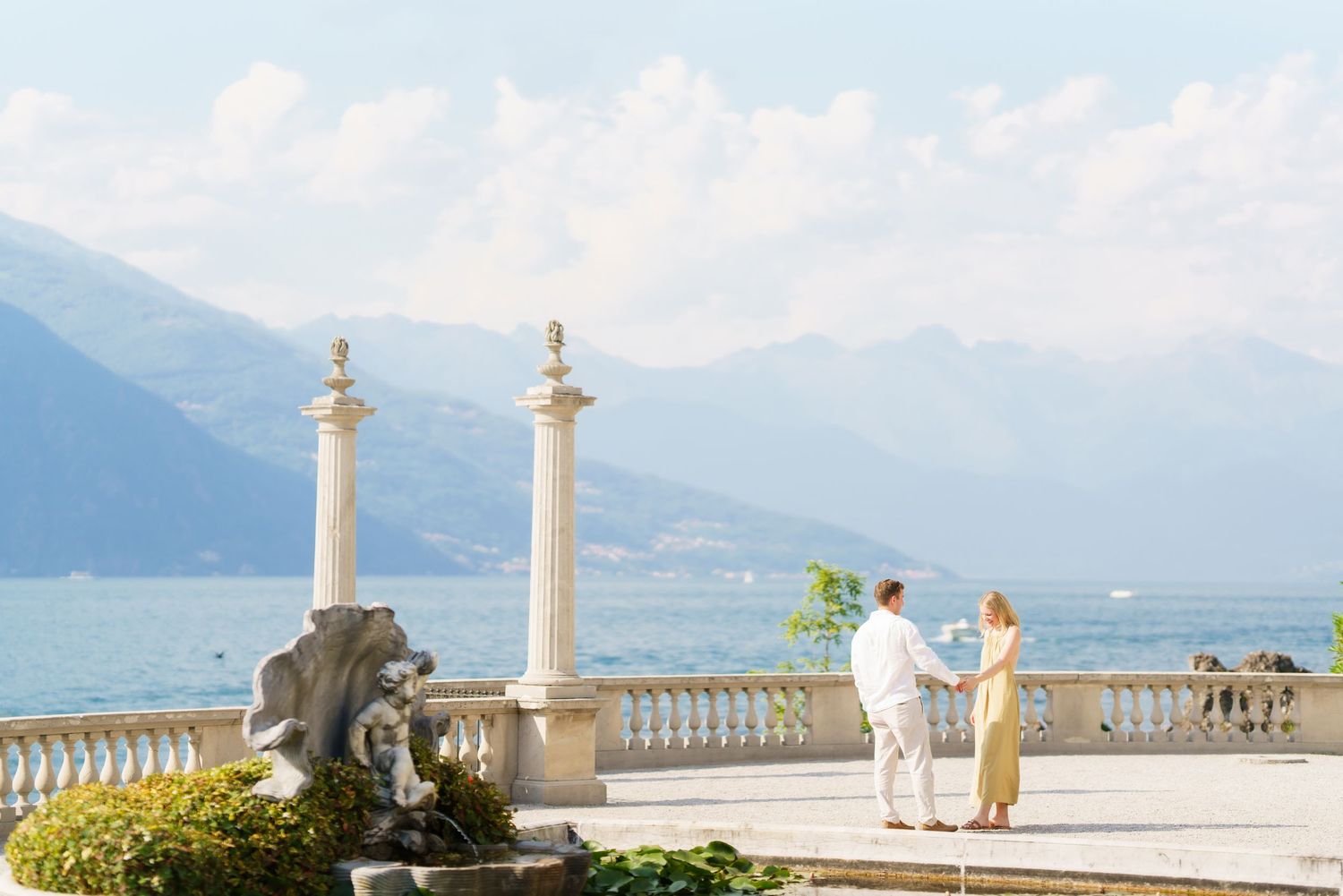 Couple standing on elegant lakeside terrace with marble columns, fountain, and mountain backdrop at Lake Como, Italy.
