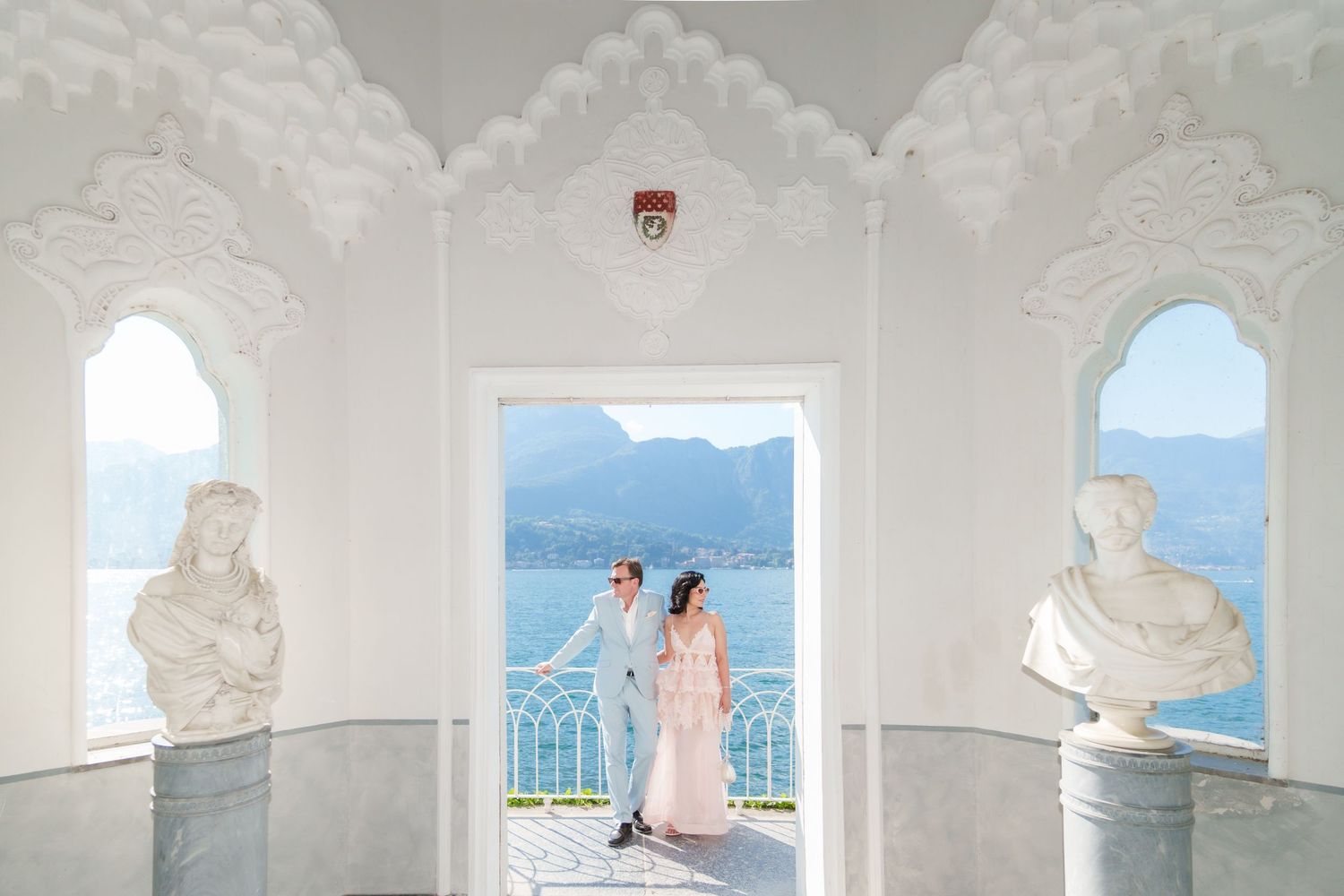 Couple in elegant attire poses in ornate white pavilion with marble busts, overlooking scenic lake and mountains.
