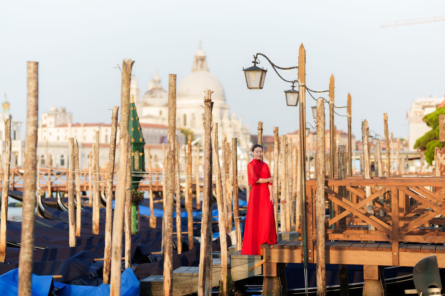 Woman in red dress standing on Venice gondola dock with historic church and lamp post backdrop.