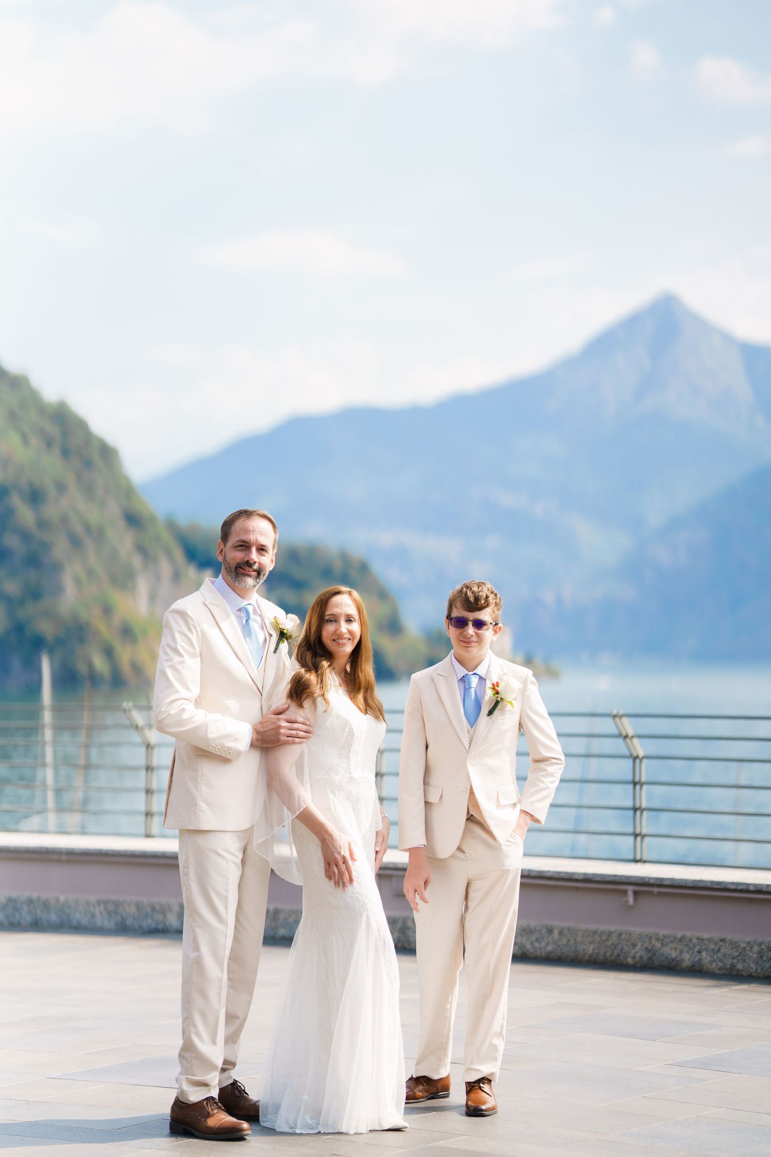Bride and groom with guest posing in cream suits by a lakeside terrace with mountain backdrop on wedding day.