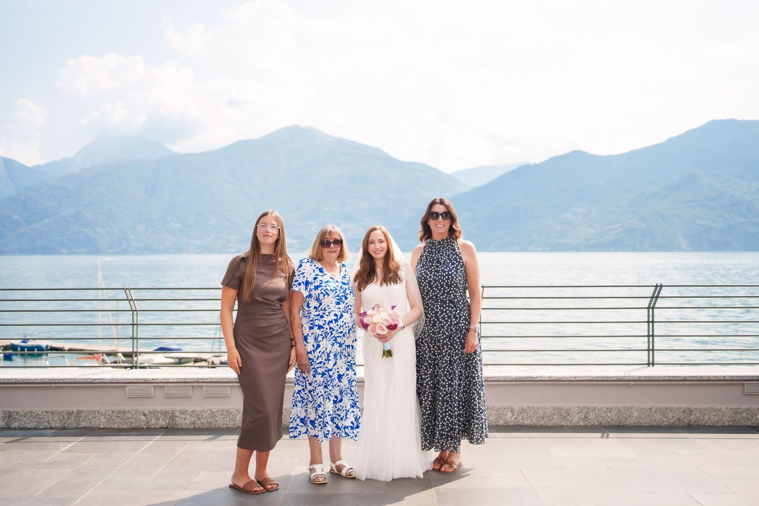 Four women pose on a lakeside terrace with mountain views, one in a white wedding dress holding a bouquet.