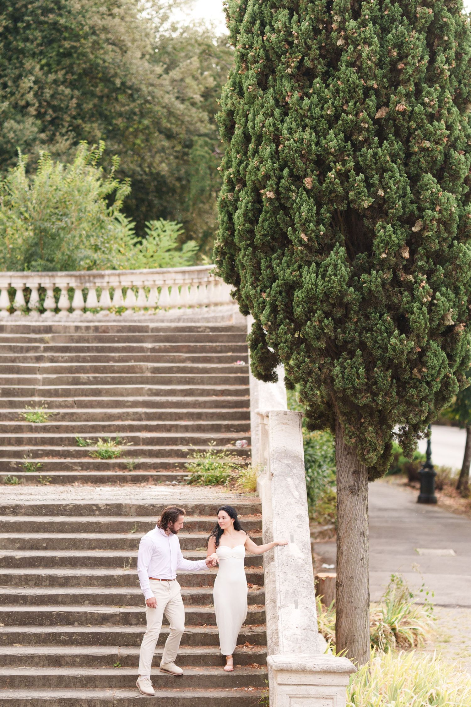 Two people in white outfits walk up stone steps in a garden setting with cypress trees.