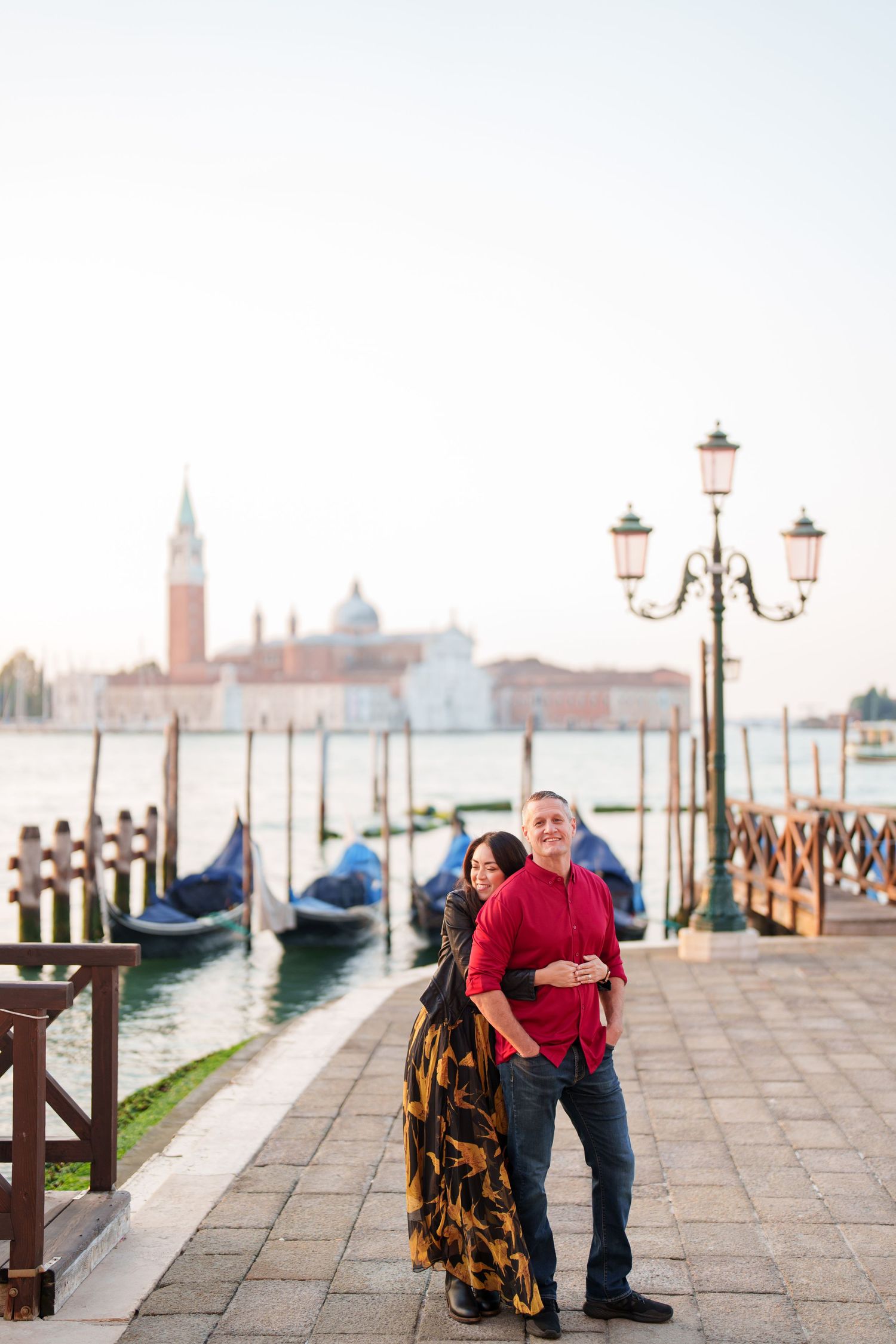 photographer venice Romantic couple embraces along Venice's waterfront with iconic gondolas and ornate lampposts in the misty background.