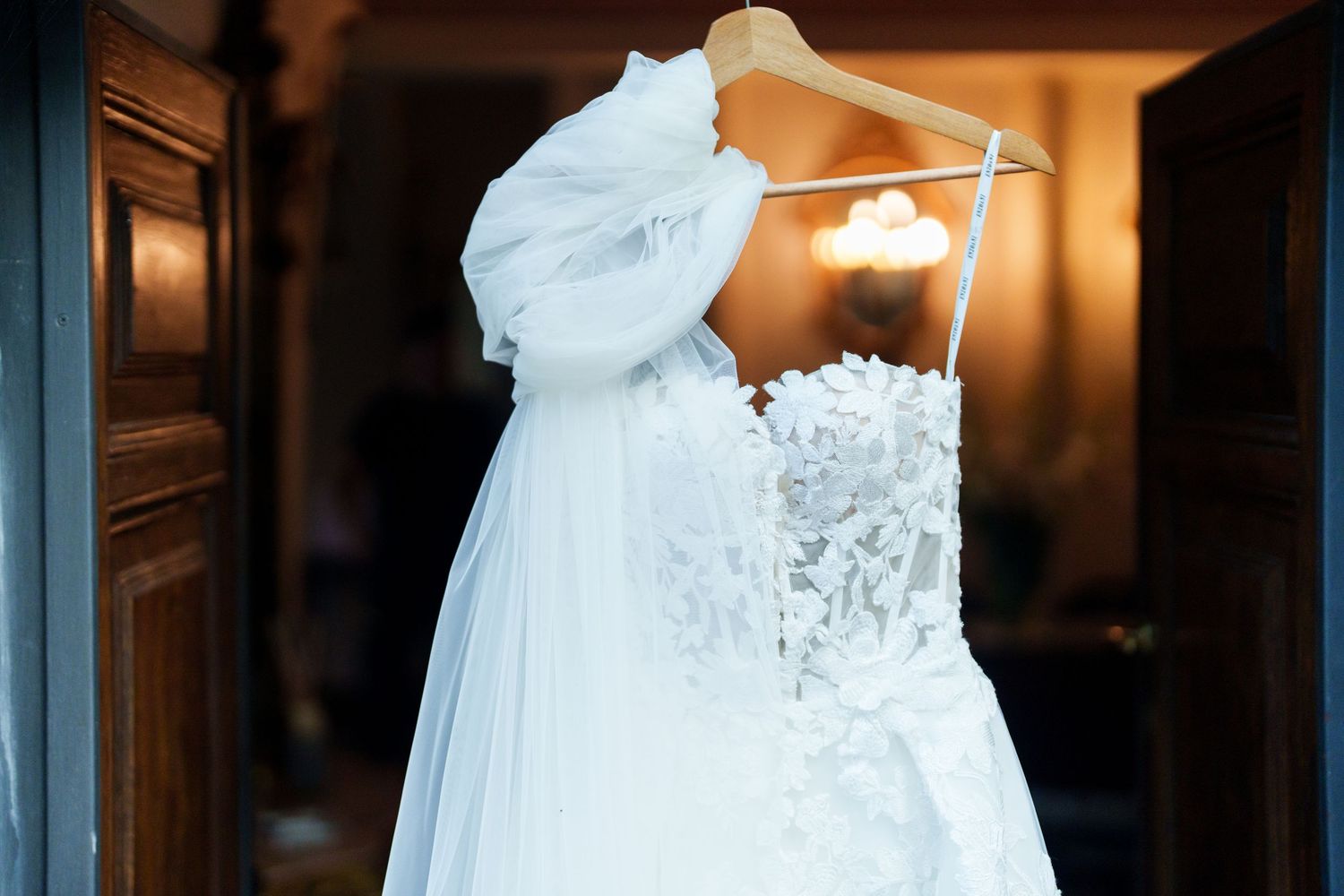 A white wedding dress with lace details hangs elegantly in front of a warmly lit doorway.