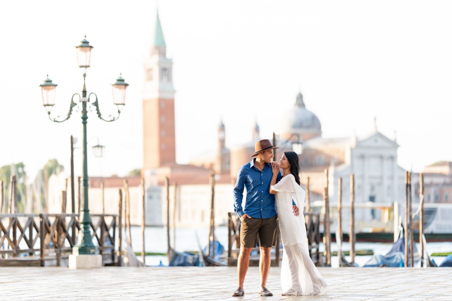 Romantic couple walking along Venice's St Mark's Square with historic architecture and lamp posts in the background.