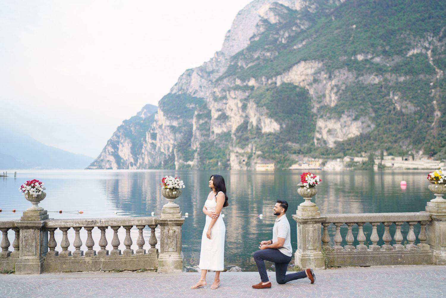 A romantic proposal moment unfolds on a stone balcony overlooking a scenic mountain lake landscape.