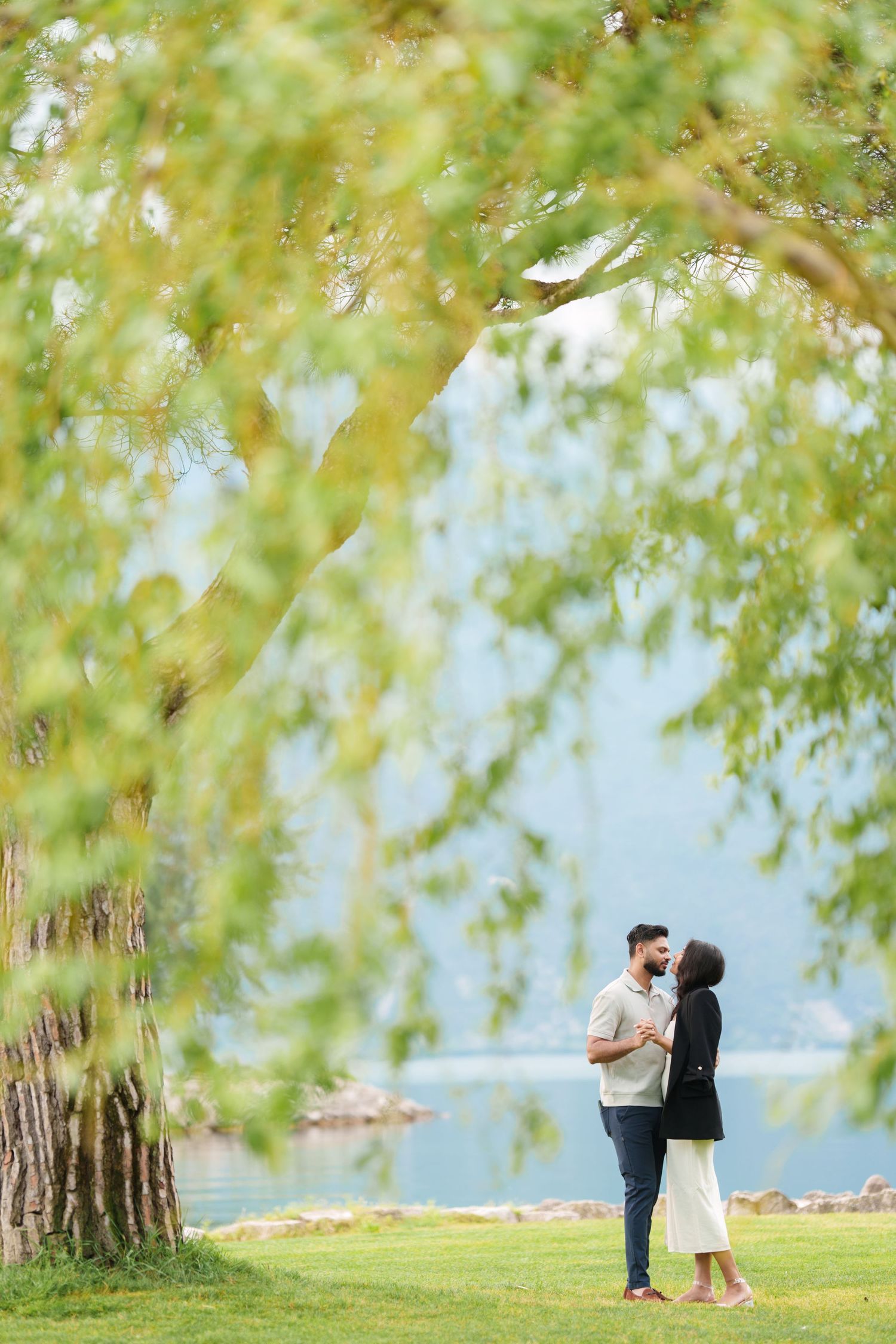 Romantic couple embraces under blooming willow trees by a serene lakeside on a bright spring afternoon.