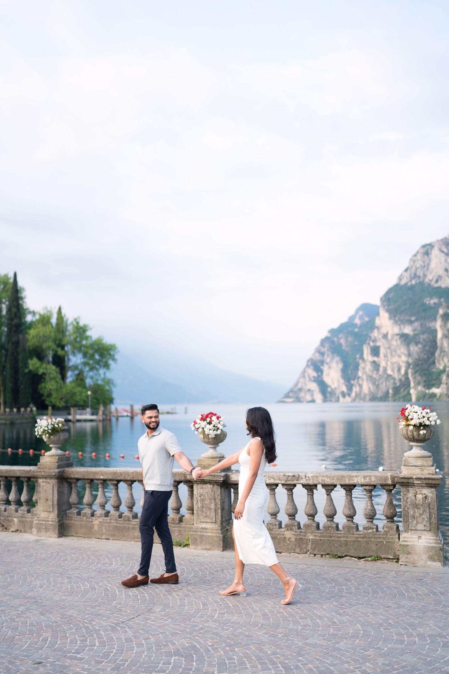A romantic couple shares a tender moment on a stone terrace overlooking Lake Garda in Italy with mountains in the background.