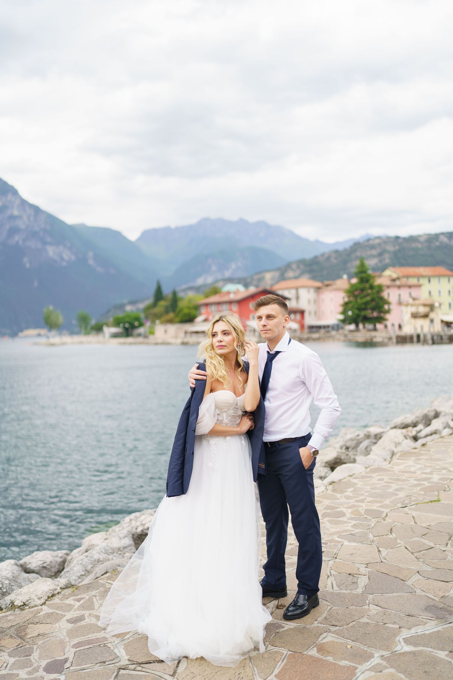 A wedding couple poses together on a stone pathway by a scenic mountain lake with buildings in the background.