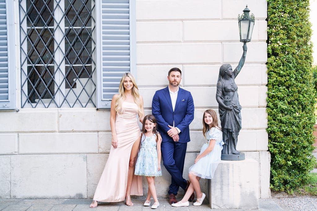 Family members in formal attire pose together outside a white stone building with vintage lamp post and lattice windows.