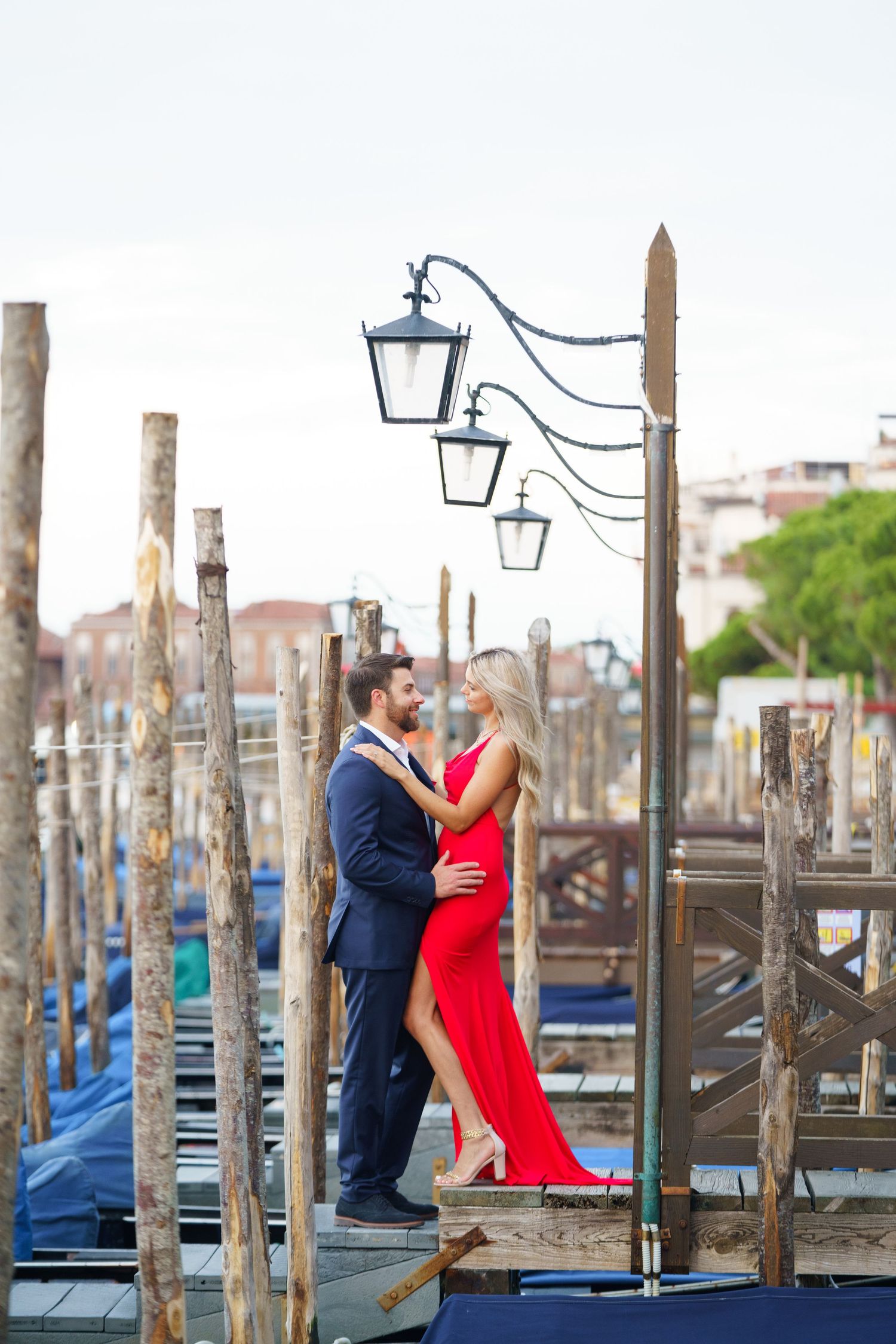 A couple in formal attire embraces romantically on a wooden dock with gondolas and lampposts in Venice.