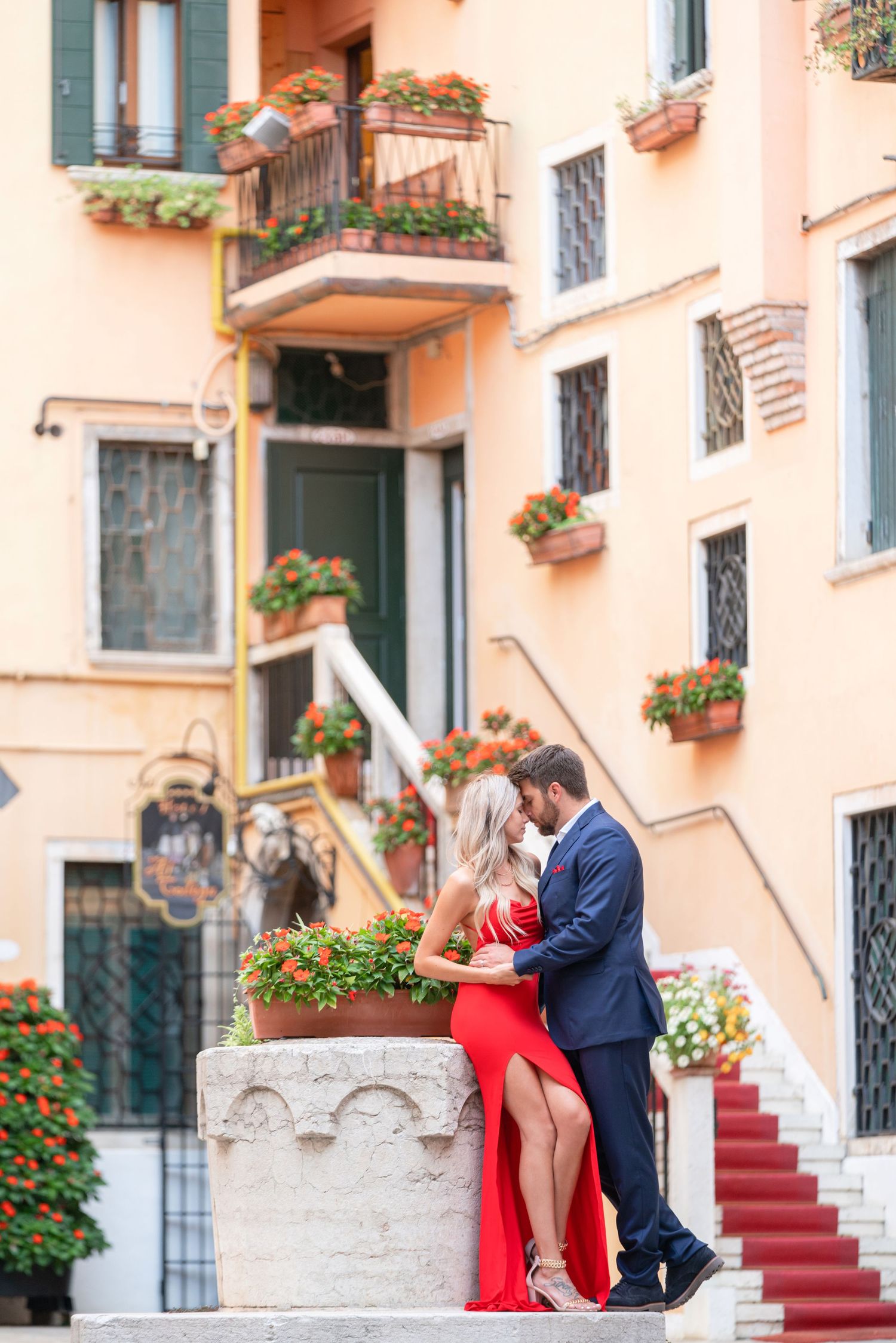 A couple in formal attire poses romantically on stone steps with red carpet and pink building facade in Venice, Italy.