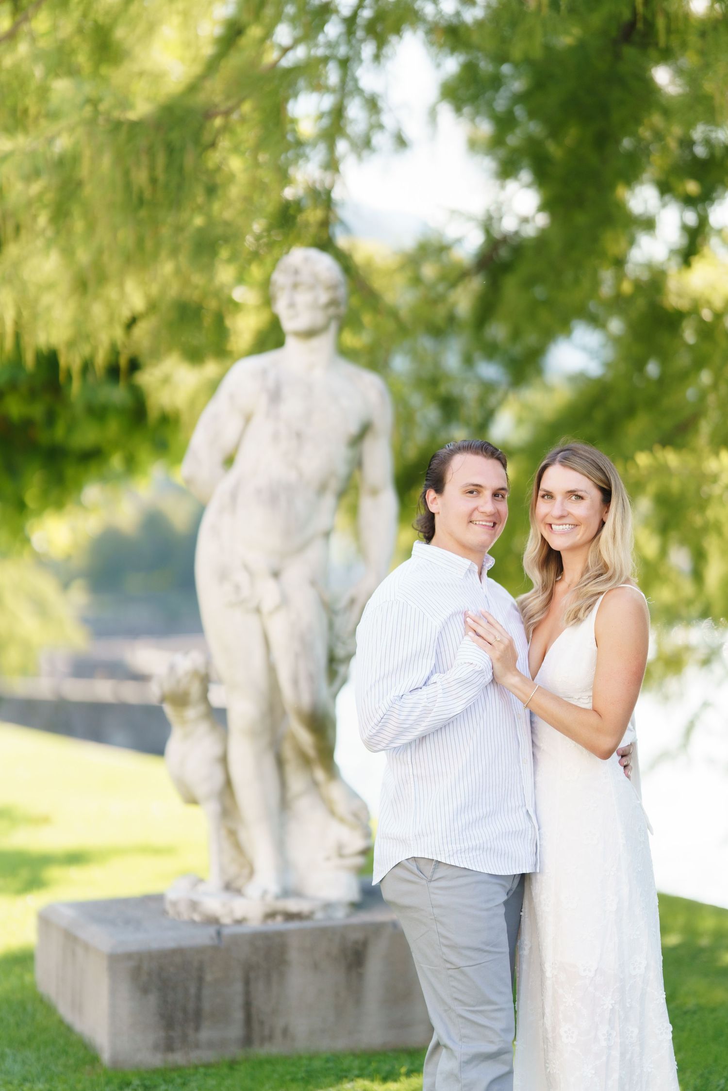 A couple shares an intimate moment in a garden setting with stone sculptures in the background.