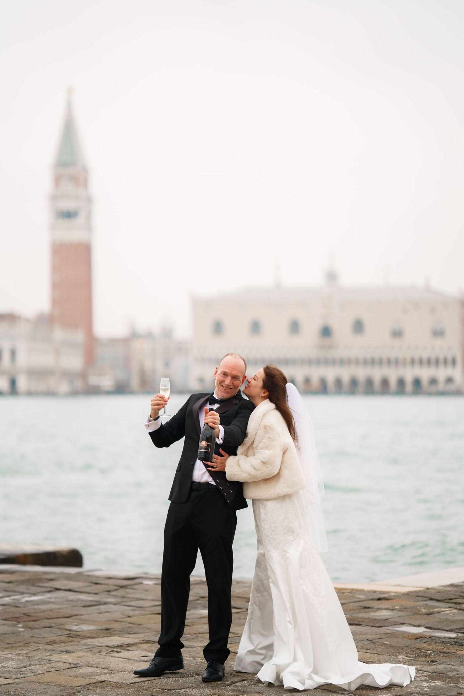 A couple in wedding attire poses along the Venice waterfront with St Mark's Campanile tower in the background.