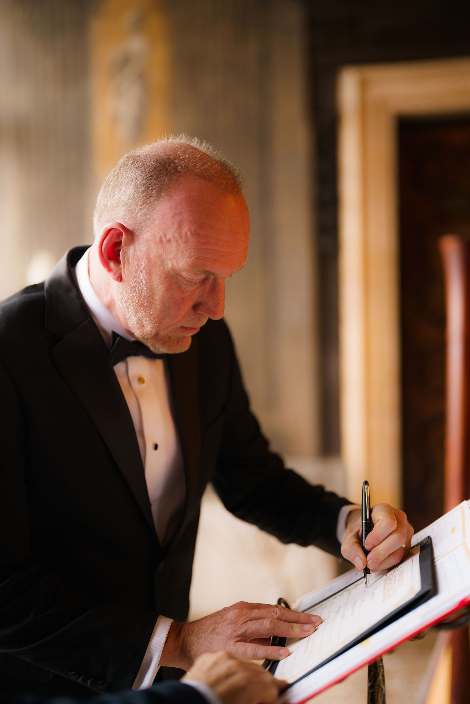 A person in formal attire signs documents in a dimly lit hallway setting.