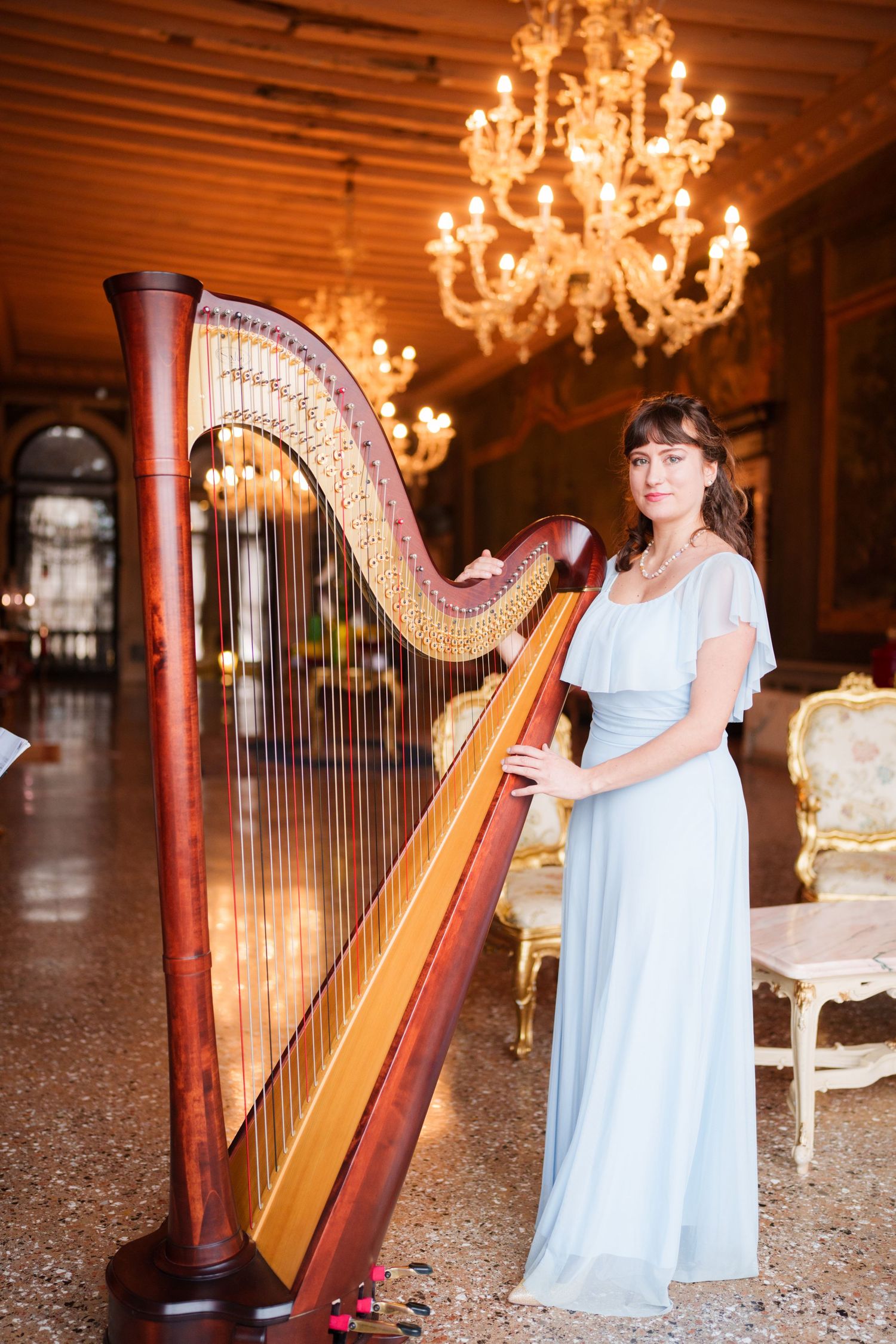 A harpist in a light blue dress performs in an ornate room with chandeliers overhead.