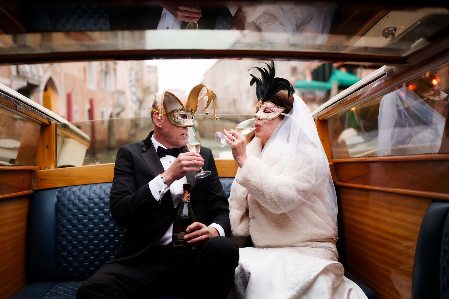 A couple in masquerade masks and wedding attire share a drink while sitting in an elegant vintage boat interior.