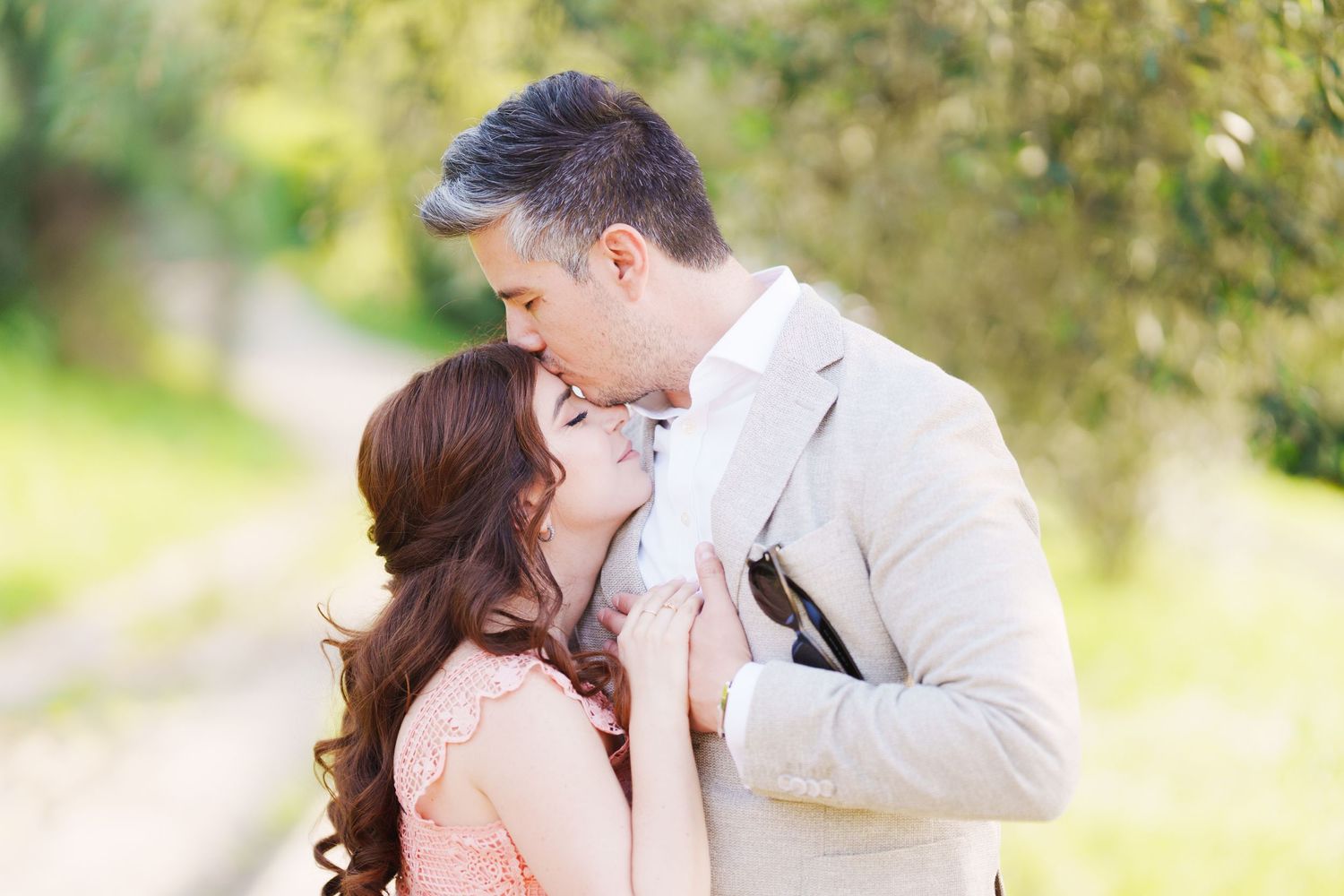 A romantic couple shares a tender kiss outdoors in soft natural lighting with blurred greenery in the background.