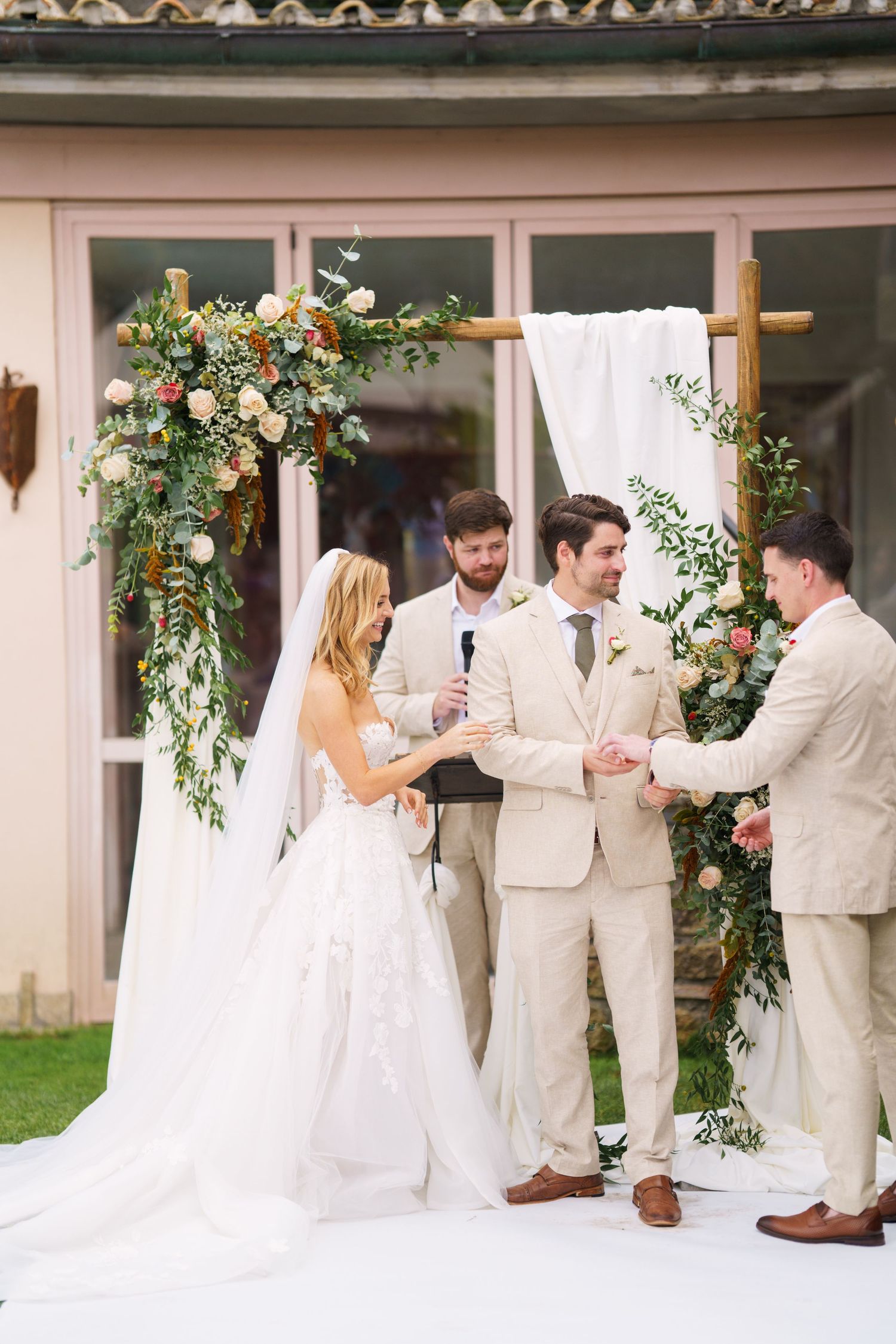 Wedding party celebrates during ceremony under an arch adorned with pink and white flowers.