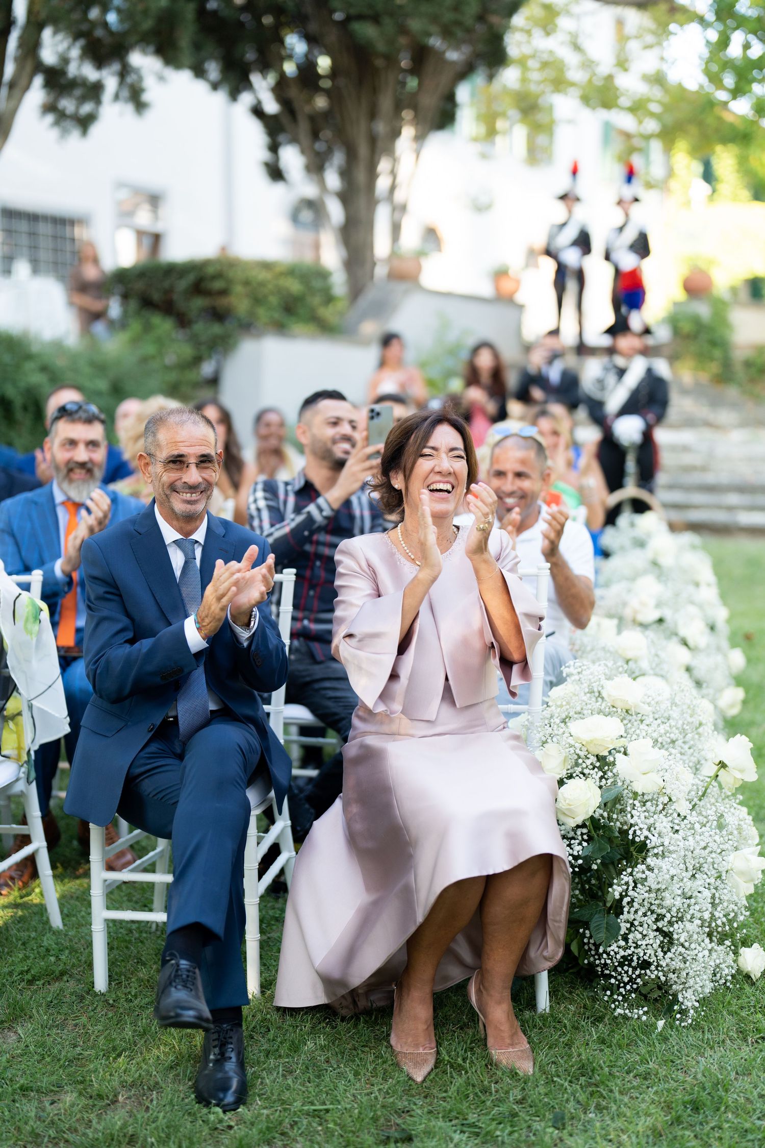 Wedding guests seated outdoors clap and celebrate during garden ceremony.