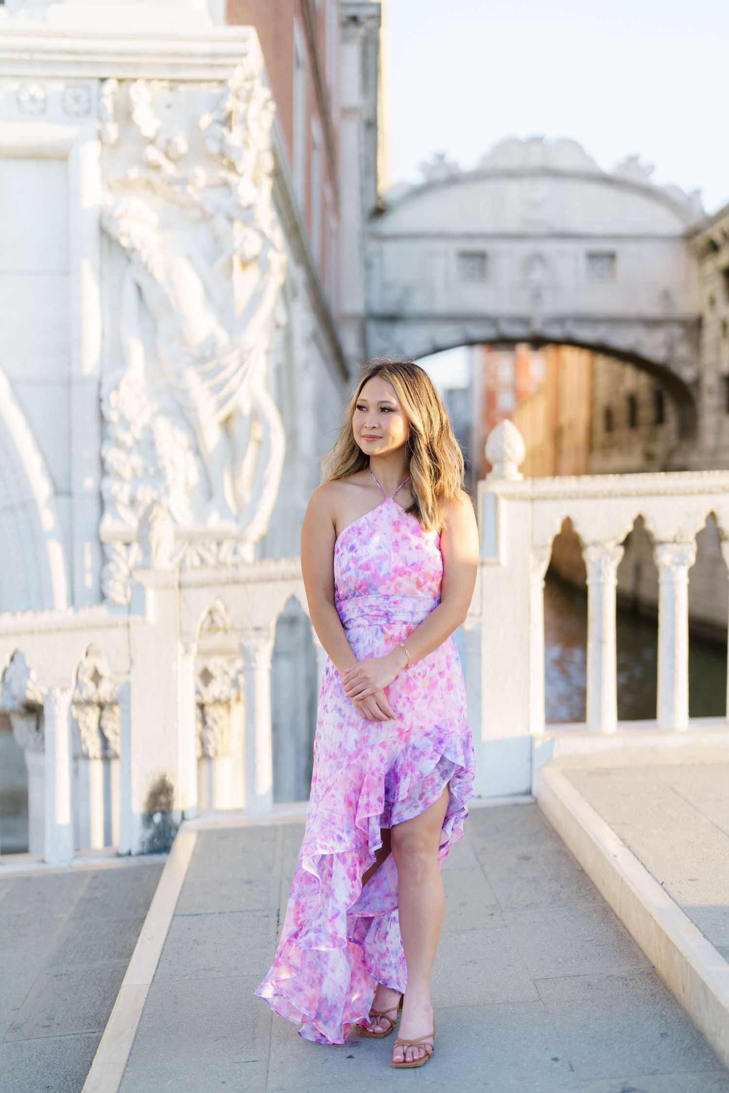 Young woman in floral pink maxi dress posing on ornate white terrace with Bridge of Sighs in background.