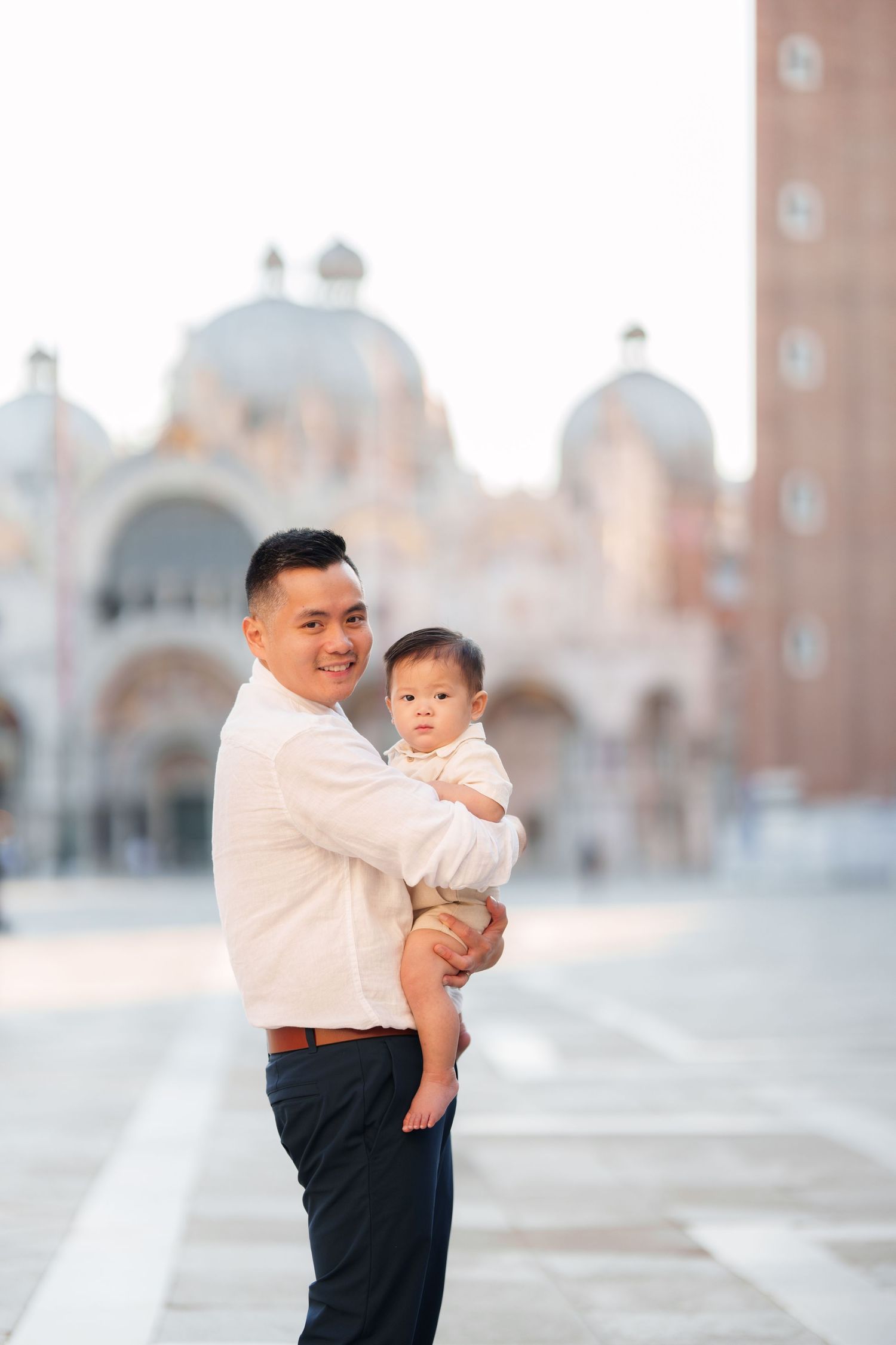 Father holding baby in St. Mark's Square, Venice, smiling at camera with Basilica di San Marco in background.