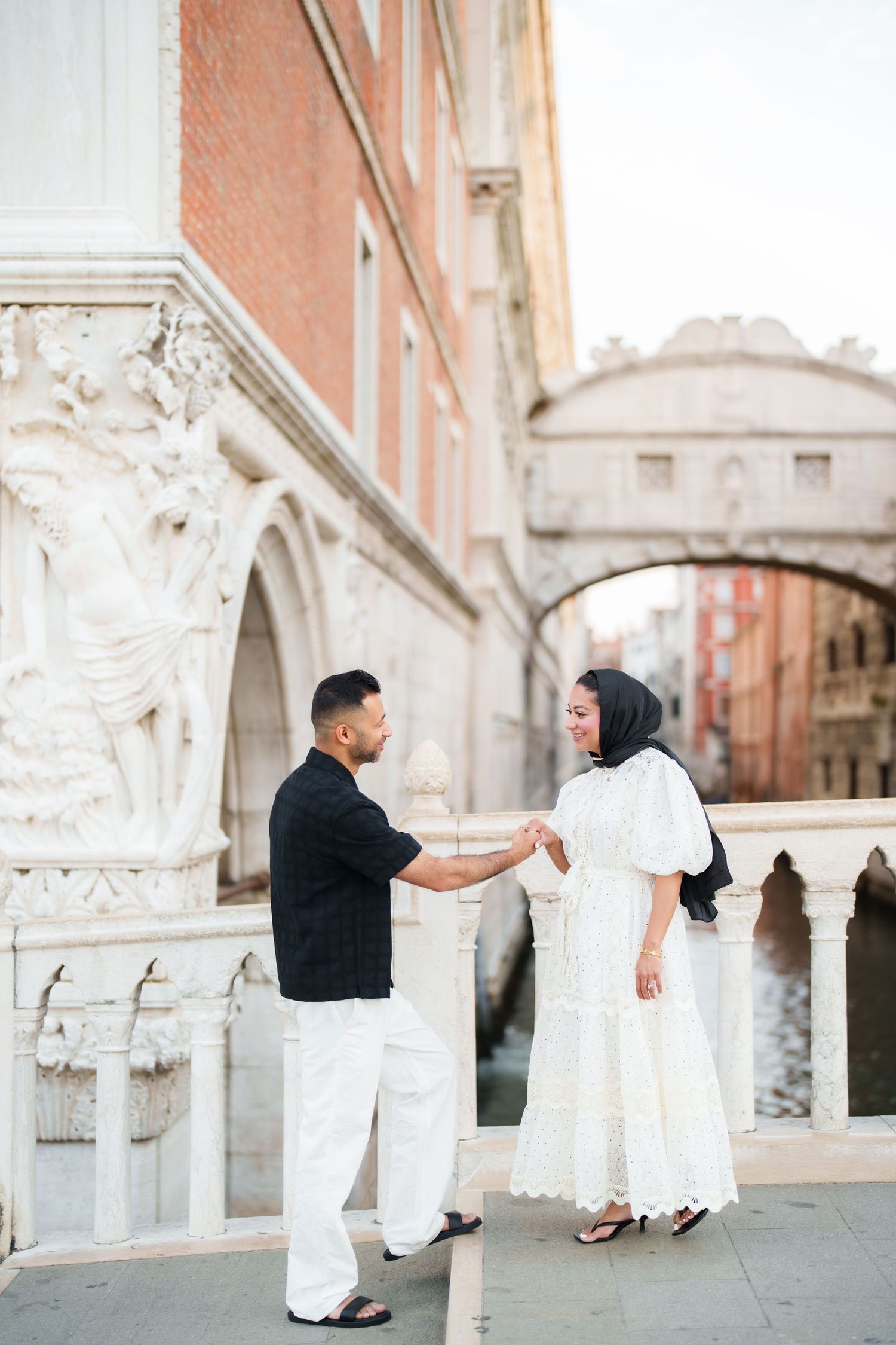 Couple holding hands near the Bridge of Sighs in Venice, woman in white dress and hijab, man in black shirt.