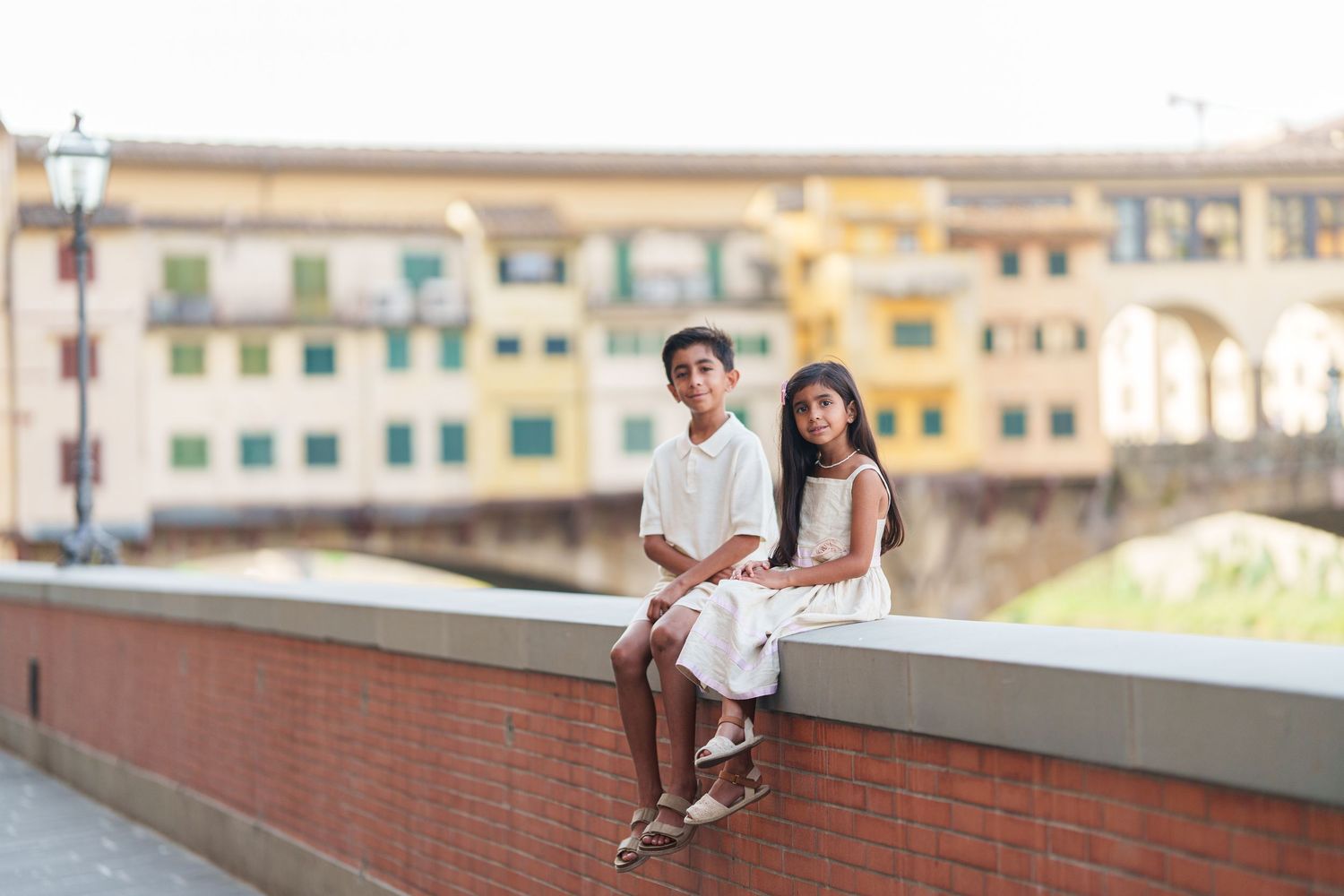 Two children in white outfits sit on a wall with Ponte Vecchio bridge in Florence, Italy in the background.