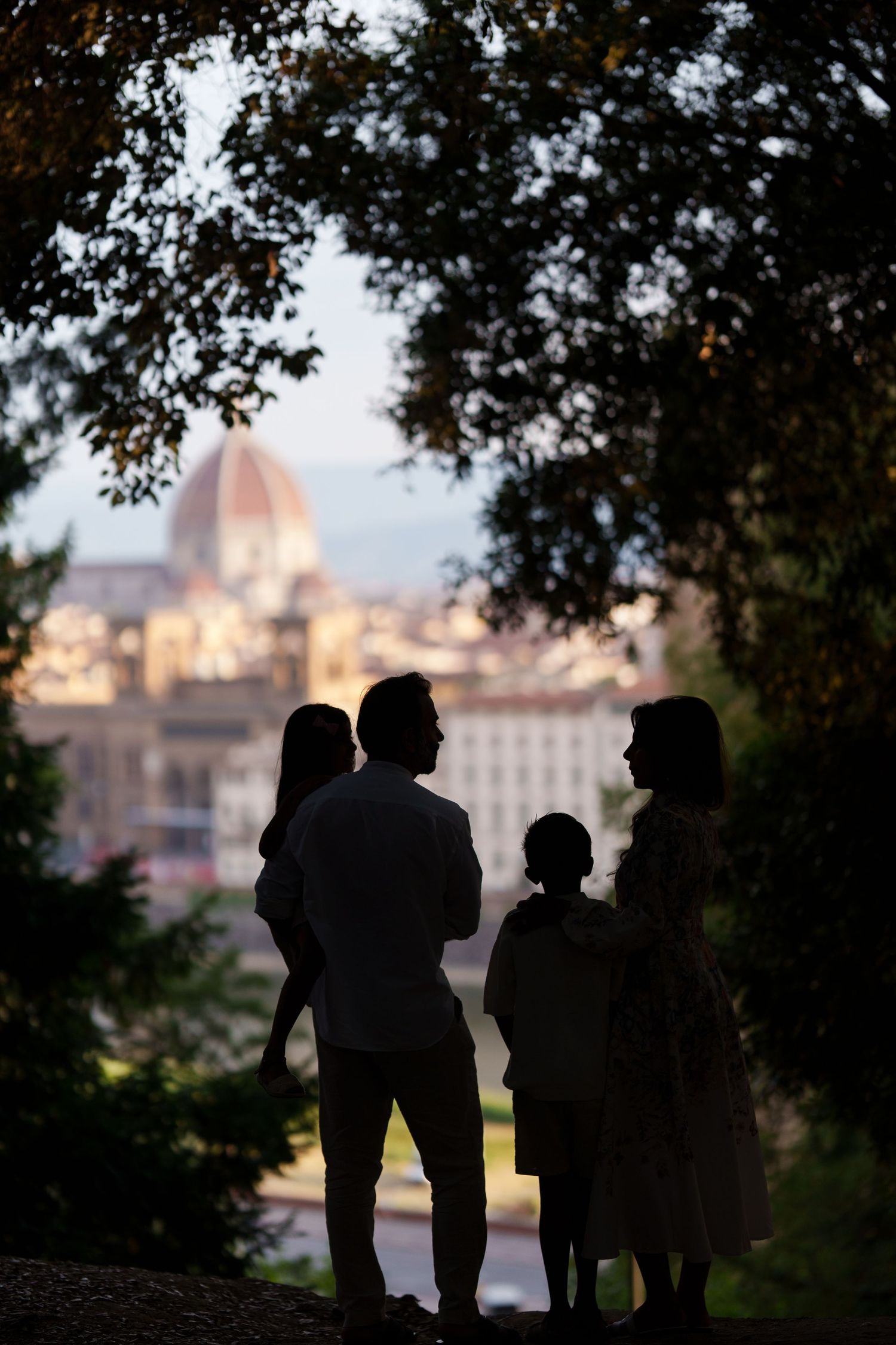 Silhouette of a family of four standing among trees with Florence cathedral dome visible in background at dusk.