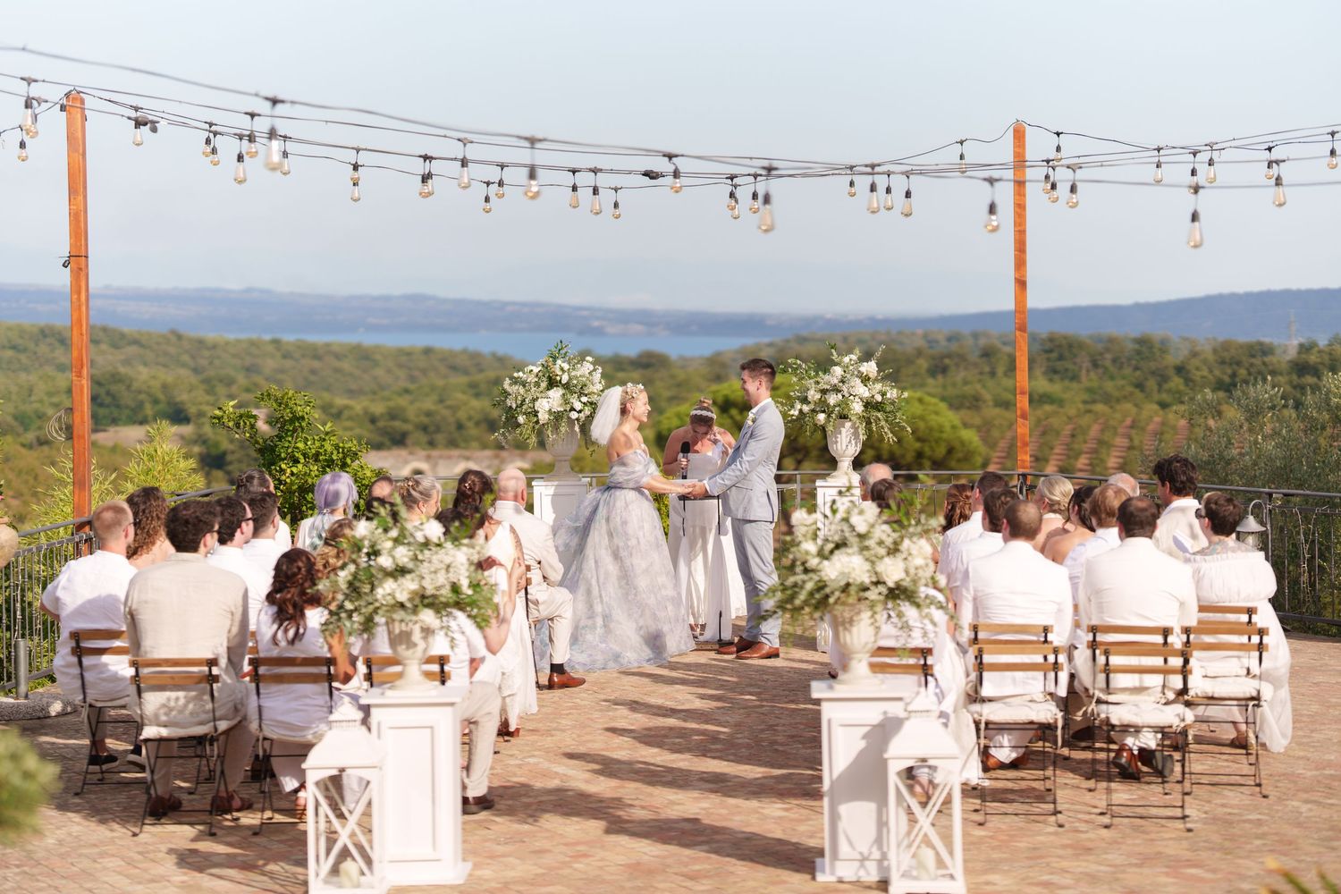 Outdoor wedding ceremony with guests in white attire, floral arrangements, string lights, and scenic forest and lake views.