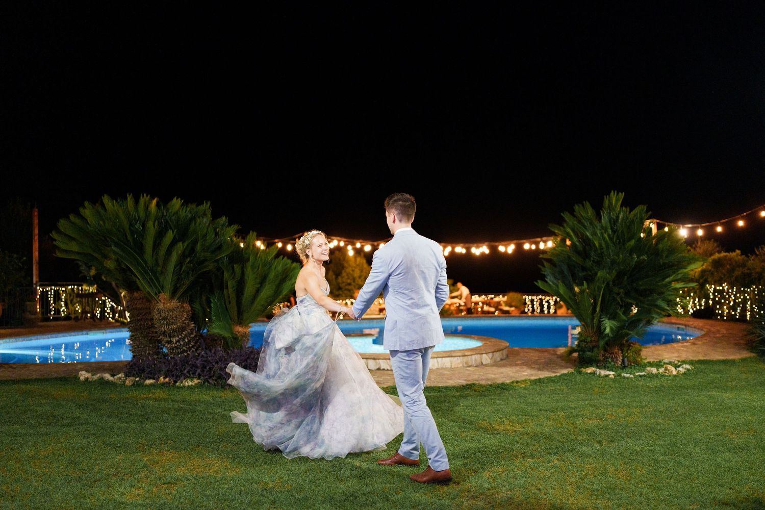 Bride and groom dancing outdoors at night near illuminated pool with string lights at elegant wedding reception.