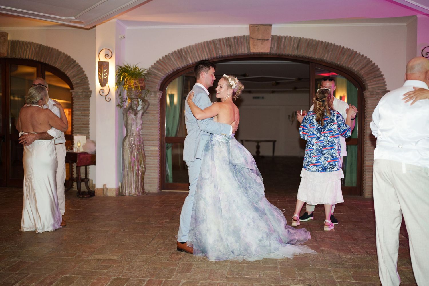 Bride and groom share first dance at elegant wedding reception venue with arched doorways and guests celebrating.