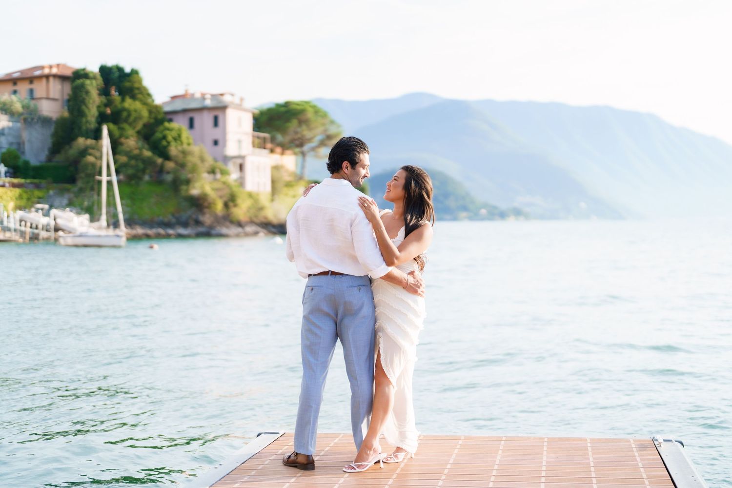 Couple embracing on a dock with scenic lake and mountain views in the background on a sunny day.