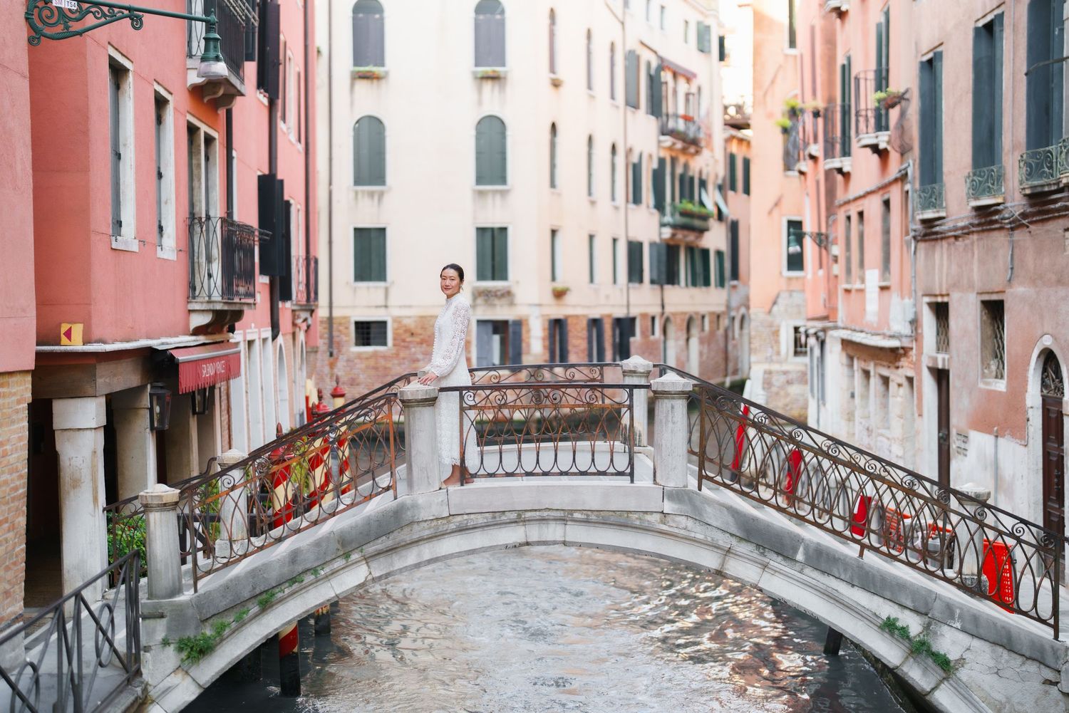 A person stands on an ornate stone bridge over a narrow Venetian canal, surrounded by colorful historic buildings.