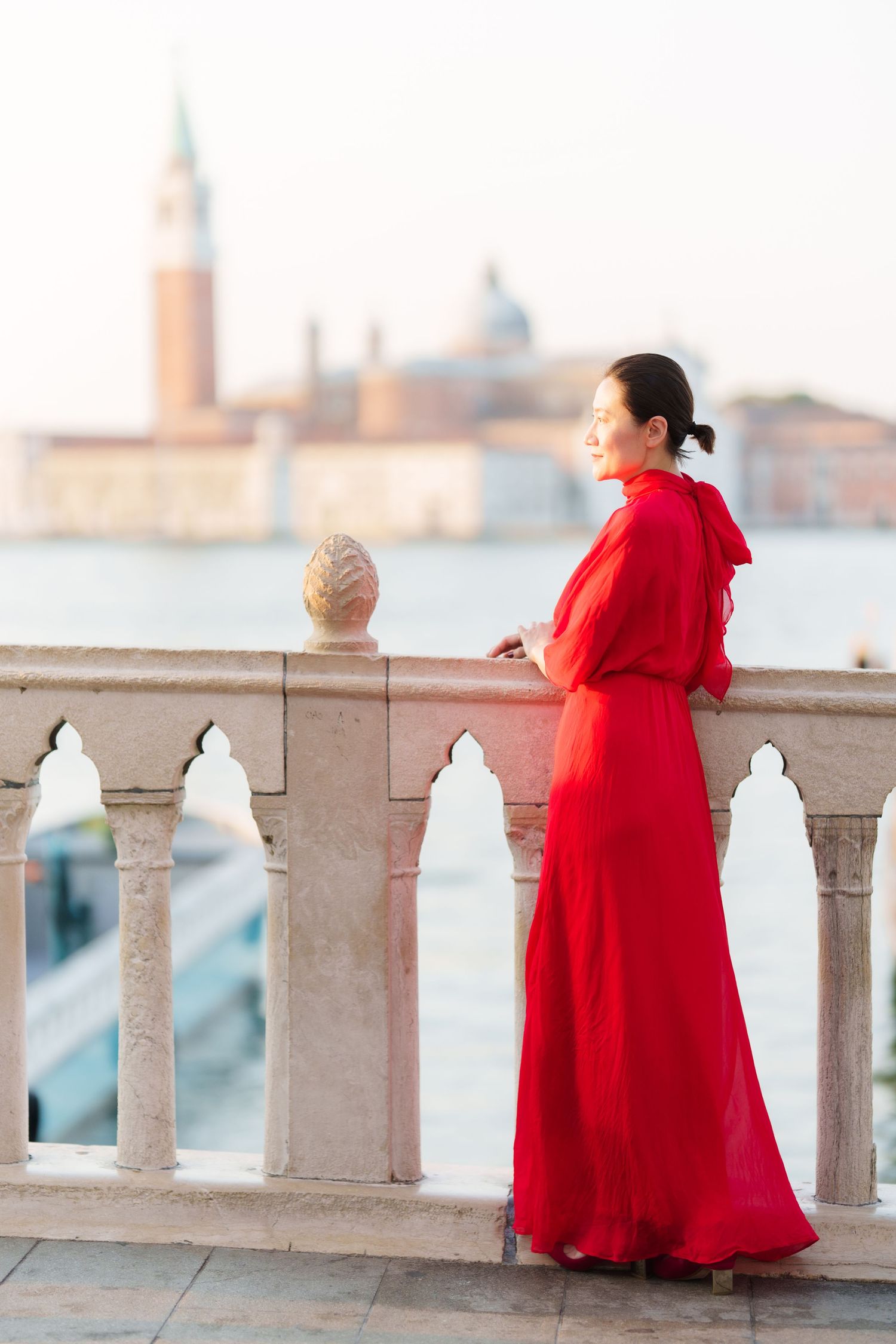 Woman in red dress standing on Venice balcony overlooking the Grand Canal with San Giorgio Maggiore in background.