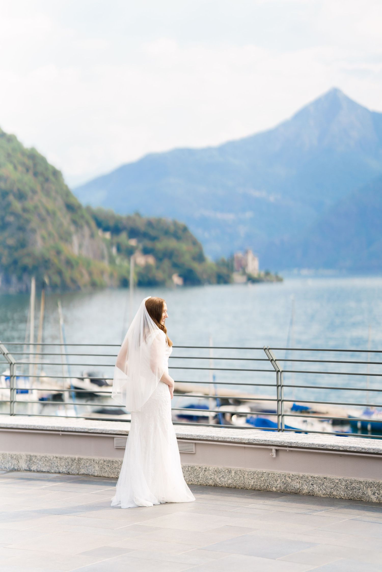 Bride in white gown and veil stands on lakeside terrace with mountain backdrop and marina views.